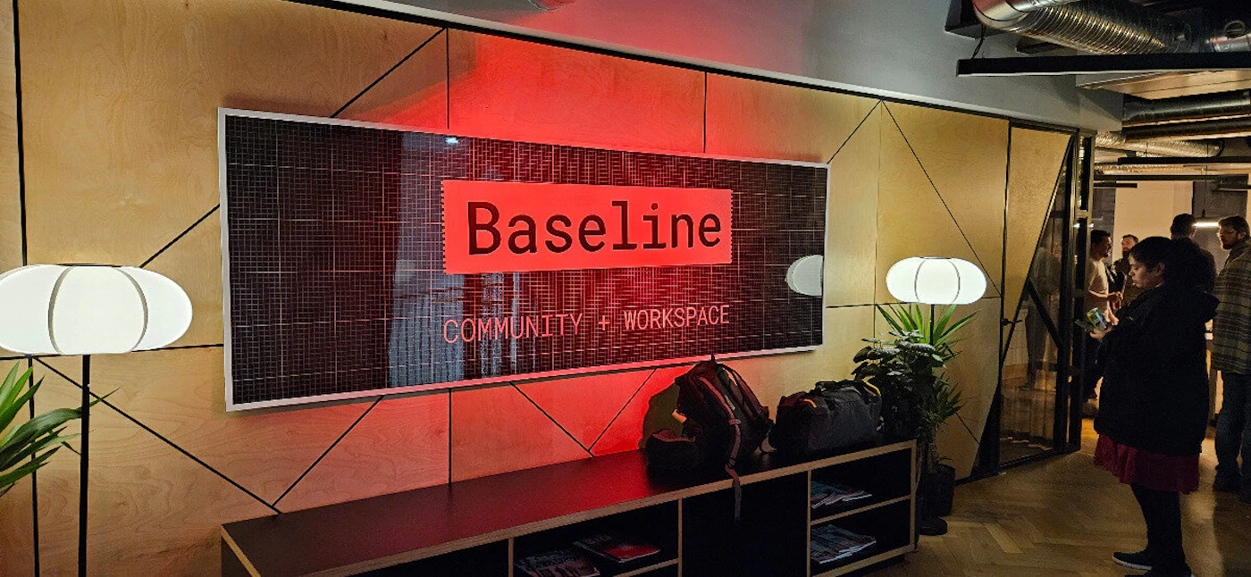 (Photo) A photo of a Baseline Community + Workspace sign inside a building, with people visible in the background. Text: Baseline COMMUNITY + WORKSPACE sign | indoor | people, interior design | candid | Colors: #DC143C, #F5DEB3, #000000 Note: The image is a real-life photograph of a sign within an office-like setting.  It captures a moment in time and is not a drawn illustration, logo, or banner in itself.