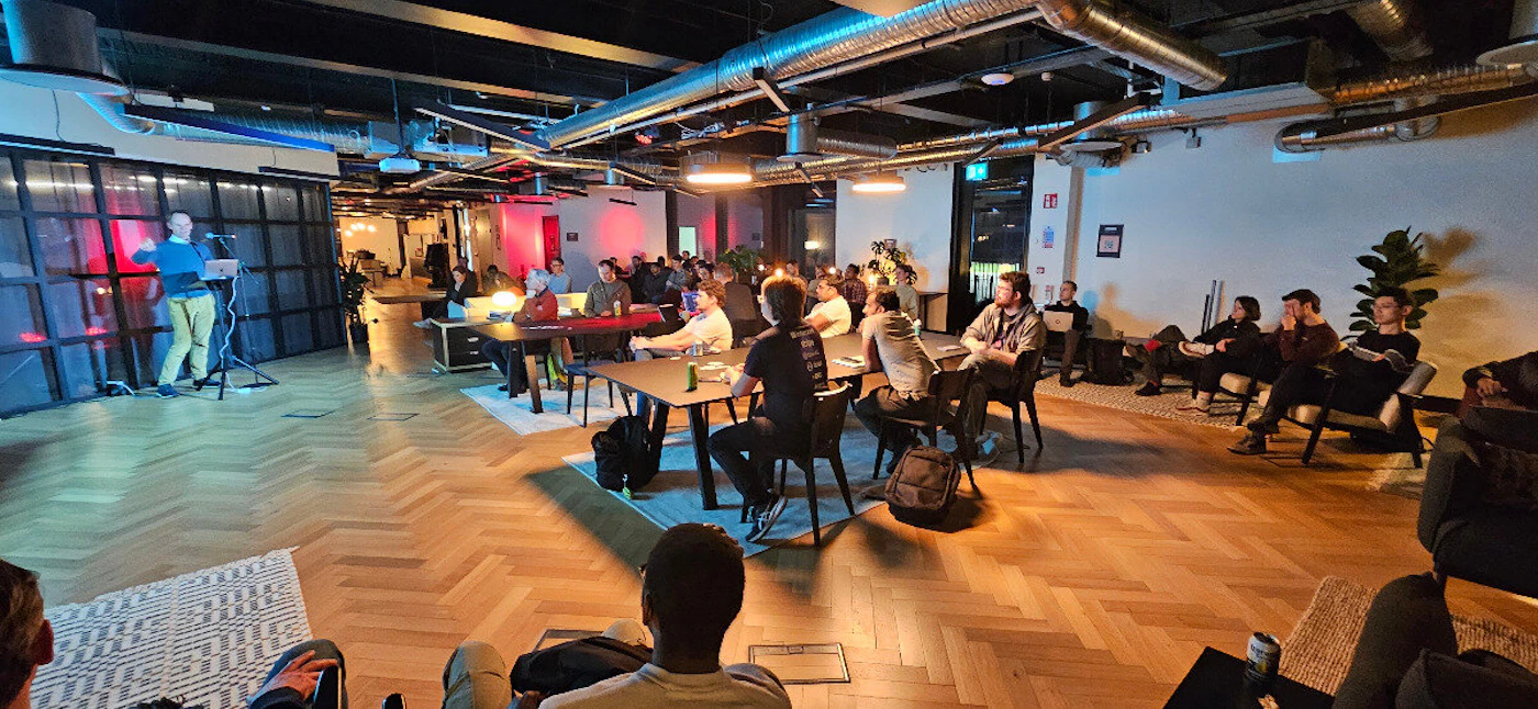 (Photo) A man gives a presentation to a group of people seated at tables and in lounge chairs in a modern office space. people | indoor | presentation, office setting, audience | candid Note: The image is a real-life captured scene of people in an office environment, making it a photograph.