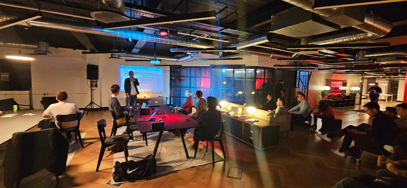 (Photo) A group of people are gathered in a modern office space, some seated around a table and others on a bench, apparently listening to a presentation projected on a screen. people | indoor | presentation, office setting, group of people | candid Note: The image is a real-life captured scene of people in an office environment, indicative of a photograph. It's not a drawn or digitally created image, nor a logo or banner.
