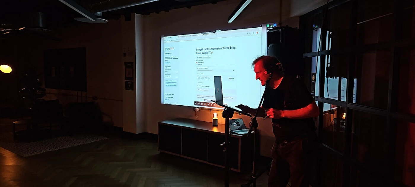 (Photo) A man is giving a presentation in a dimly lit room. A screen displays a webpage with text on it. Text: BlogWizard: Create structured blog from audio person | indoor | presentation, screen display | candid | Colors: #333333, #FFFFFF, #AAAAAA Note: The image shows a real-world scene with a person and objects, suggesting it's a photograph rather than an illustration or logo. There is text visible on the screen.