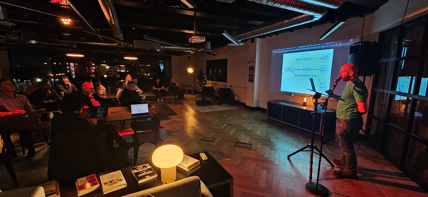 (Photo) The image shows a man giving a presentation to a group of people in a room. The presenter is standing next to a screen displaying a diagram. person | indoor | multiple people, presentation screen | candid | Colors: #282828, #BDBDBD, #F5F5DC Note: The image is a realistic depiction of a scene, suggesting it is a photograph rather than an illustration or other type of image.