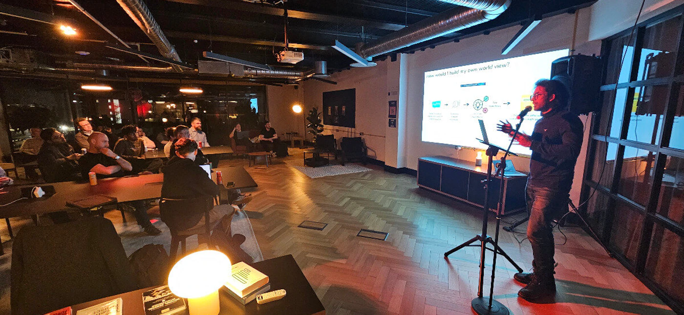 (Photo) The image shows a presentation being given to an audience in a room with a projector screen. A man is standing at the front, presenting to a group of people seated at tables. Text: How would I build my own world view? communication people, presentation | indoor | projector screen, audience, presenter | candid | Colors: #282828, #FFFFFF, #A07A5F Note: The image shows a real-world scene with people and objects captured by a camera, making it a photograph. The presence of an audience and a presenter suggests a real event.