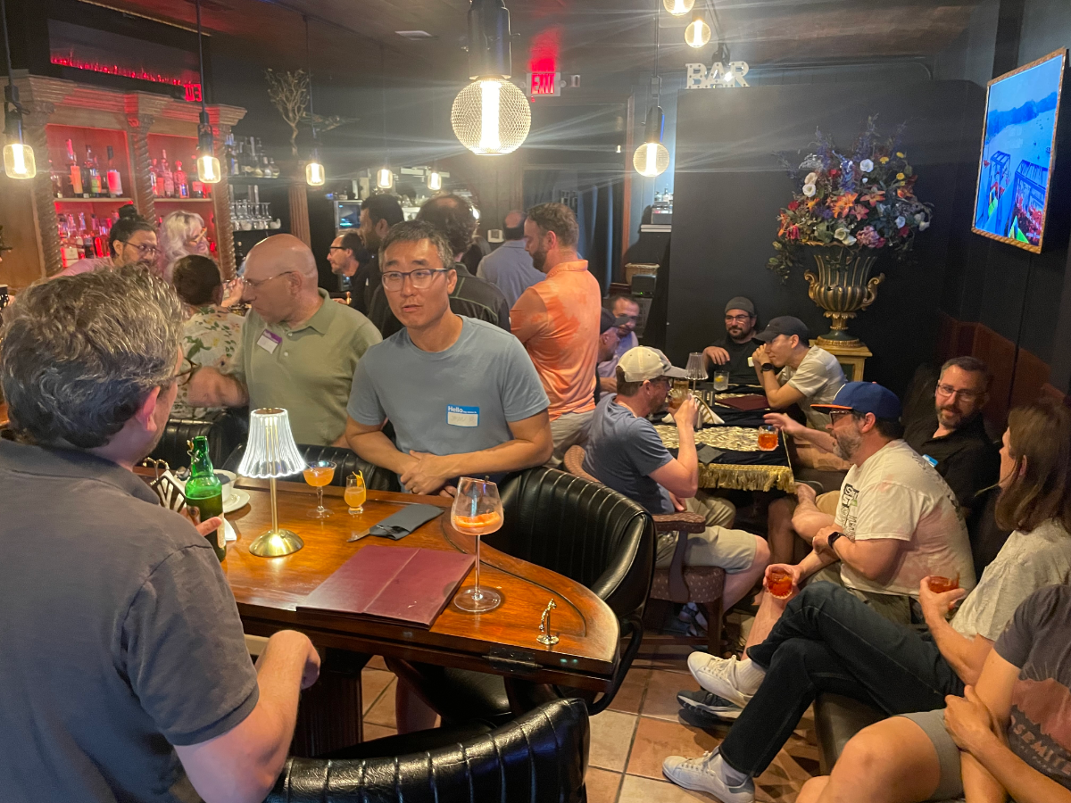 (Photo) A candid indoor photograph captures a diverse group of people socializing in a dimly lit bar or lounge, with some standing at the bar counter and others seated at various tables. Text: EXIT BAR Hello People socializing at a bar/lounge | Indoor, a dimly lit bar or lounge | Multiple individuals engaged in conversation, a well-stocked bar, various drinks on tables, and decorative elements like a large floral arrangement and framed art. | #222222 | #a06020 | #6a8094 | #8b9d7a | #ff0000 | Candid, event photography, warm and dim lighting | Colors: #222222, #a06020, #6a8094, #8b9d7a, #ff0000 Note: The image is a direct, unedited capture of a real-world scene, depicting people and an environment, which is characteristic of a photograph.