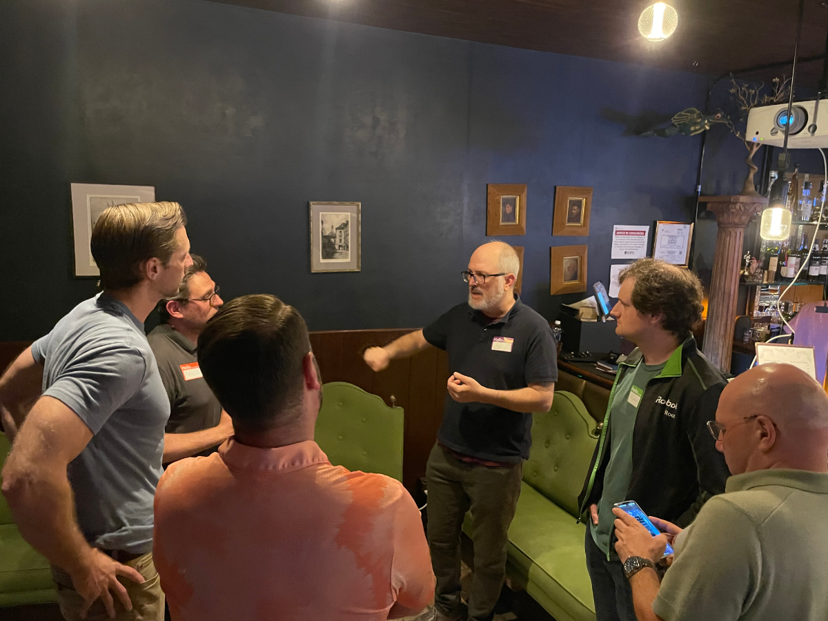 (Photo) A group of men are gathered in a dimly lit indoor setting, likely a lounge or bar, engaged in conversation. One man in the center is actively speaking and gesturing to the others. Text: Rebol A group of men socializing | Indoor, a lounge or bar area with dark walls and vintage-style furniture. | Multiple people (at least 6 visible), conversational interaction, dim and warm lighting, framed pictures on the wall, a bar counter in the background. | #263640 | #658B43 | #E09060 | #90A0B0 | #F0E68C | Candid, informal group shot | Colors: #263640, #658B43, #E09060, #90A0B0, #F0E68C Note: The image captures a real-life scene featuring multiple individuals and their environment, characteristic of a photograph rather than a stylized illustration, logo, or text-focused banner.