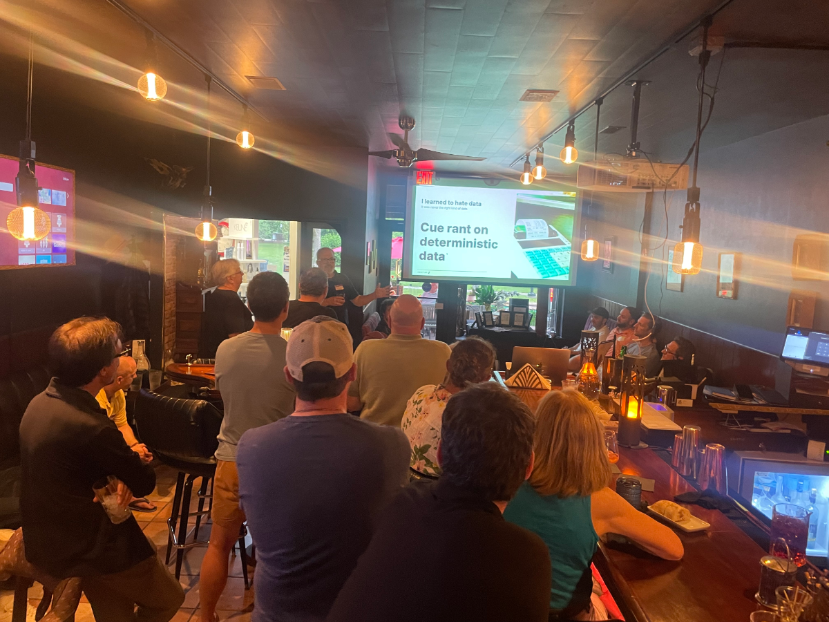 (Photo) A candid indoor photograph shows a group of people gathered in a dimly lit bar or pub, facing a projector screen that displays a presentation slide. The room is illuminated by warm, hanging light fixtures. Text: I learned to hate data and the spreadsheet life Cue rant on deterministic data People attending a presentation or social gathering | Indoor, likely a bar or pub with a projector setup | Group of individuals, projector screen with text and an image, bar counter, ambient lighting from hanging bulbs, some people are seated at the bar while others stand. | #2E2E2E | #F8A800 | #808080 | #4A8D6D | #504F47 | Candid, event photography, low light | Colors: #2E2E2E, #F8A800, #808080, #4A8D6D, #504F47 Note: The image is a direct capture of a real-world scene, featuring people and an environment, which aligns with the definition of a 'photo'.