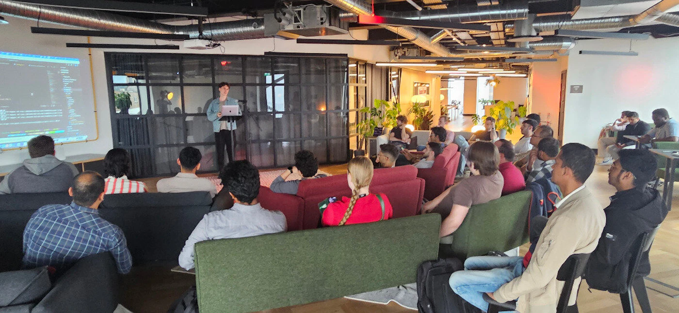 (Photo) A photograph showing a technical presentation or workshop held in a modern office space with an industrial aesthetic. Text: [unreadable] Group of people attending a presentation/workshop | Indoor, modern office or co-working space with exposed ceiling ducts and comfortable seating. | A presenter stands near a projection screen displaying lines of code; the audience is seated on mixed-color couches (green, red, dark gray). | Candid/event photography, wide shot. | Colors: #a0846c, #333333, #5e824e, #8b0000 Note: The image is a captured moment of a real-life event involving a group of people and a specific indoor environment, classifying it as a photograph.