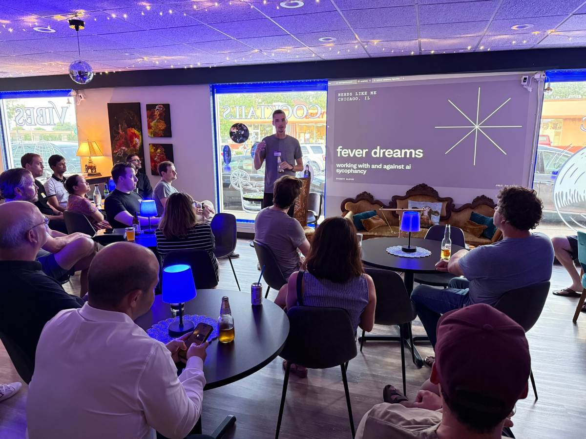 (Photo) The image captures an indoor gathering where a group of people are seated, facing a speaker who is presenting on a large screen. The setting appears to be a lounge or bar, characterized by ambient purple lighting, small tables with blue lamps, and a view of the street outside through a large window. Text: NEEDS LIKE ME CHICAGO, IL fever dreams working with and against ai sycophancy ROBERT SAUER VIBES COCKTAILS A group of people attending a presentation or talk. | Indoor, likely a bar or lounge, with large windows looking out onto a street. | A speaker presenting to an audience, a large screen displaying text and a graphic, ambient purple string lights, blue table lamps, a disco ball, and various seating arrangements. | #4A3B9B | #2E2E2E | #8C8C8C | #FFFFFF | #6B6B6B | Candid, event photography. | Colors: #4A3B9B, #2E2E2E, #8C8C8C, #FFFFFF, #6B6B6B Note: This image is a realistic capture of a real-world scene featuring people, objects, and a specific environment, which aligns with the definition of a 'photo'. It is not a drawing, logo, or a simple text banner.