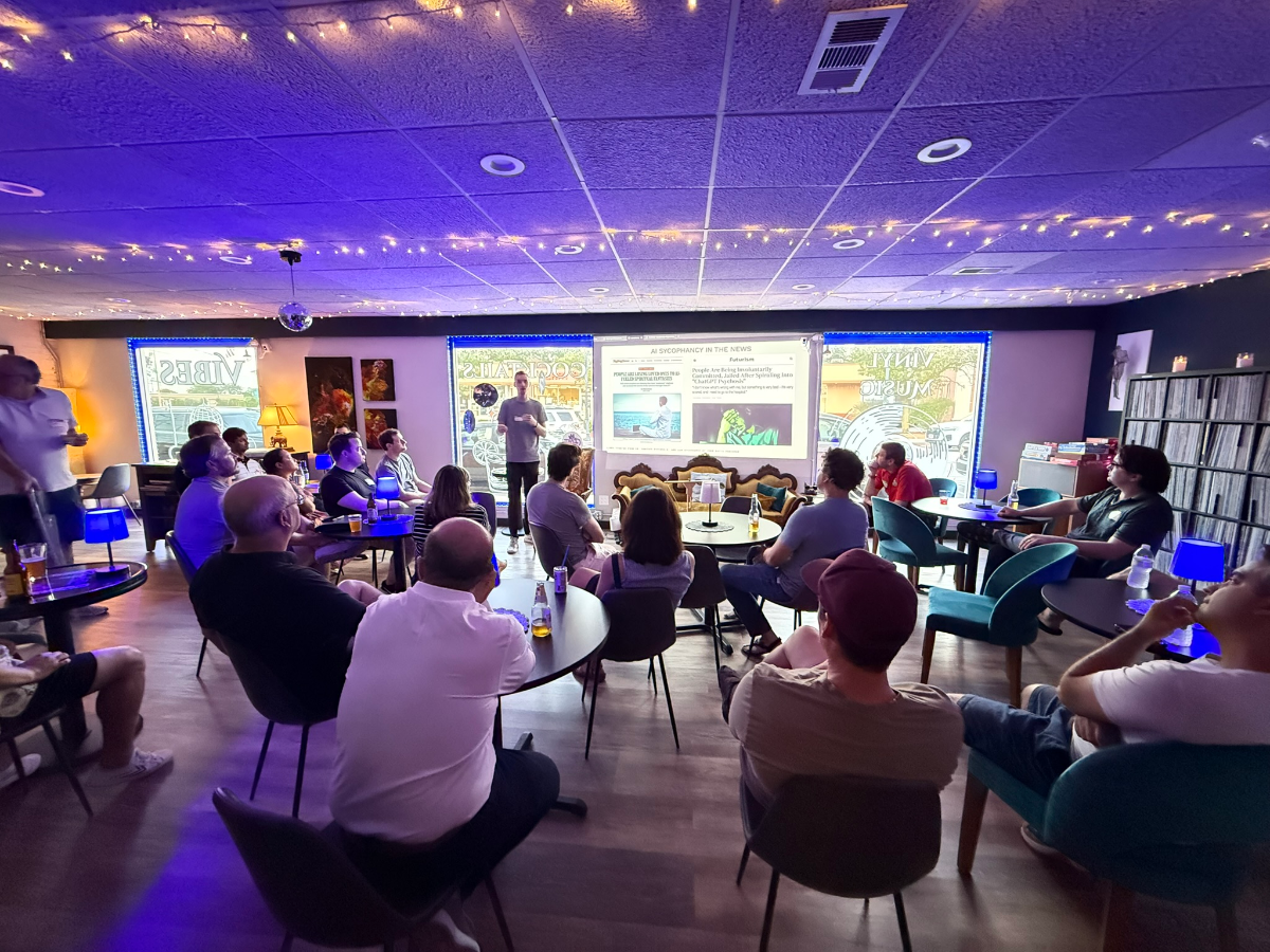 (Photo) A group of people are gathered indoors, seated around small tables, watching a presentation projected onto a screen at the front of the room. The room is dimly lit with string lights and a strong purple ambient light, and reflections of text like 'VIBES' and 'COCKTAILS VINYL MUSIC' are visible reversed on the windows. Text: AT SIGGRAPH IN THE NEWS VIBES COCKTAILS VINYL MUSIC people attending an event or presentation | indoor, likely a bar or event space | multiple people, projector screen with presentation, string lights, ambient purple lighting, record shelves, reflections on windows | #6A0DAD | #FFD700 | #2F4F4F | #008080 | candid, event photography | Colors: #6A0DAD, #FFD700, #2F4F4F, #008080 Note: This image is a direct capture of a real-world scene featuring people and an indoor environment, which clearly categorizes it as a photograph.
