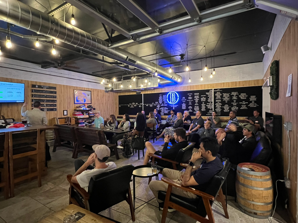 (Photo) A group of people are gathered in a casual, rustic-industrial indoor space, likely a brewery or bar, attending a presentation. A man stands at a counter, gesturing towards a screen displaying a presentation, while others are seated in chairs and benches, watching. Text: proof % works? +16% +90% VIP 1 2 3 4 5 6 7 8 9 10 B People attending a presentation or event | Indoor, a bar or event space with wooden paneling, exposed ductwork, and string lights. | Multiple people, a presenter, a screen displaying data, a neon 'B' sign, barrels, and a bar counter. | Candid, event photography | Colors: #8B4513, #36454F, #FFD700, #0000FF, #A0522D Note: This image captures a real-world scene with multiple people and an environment using photographic techniques, rather than being an illustration, logo, banner, or handwriting.