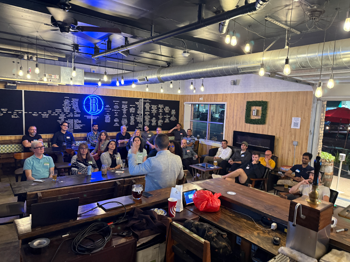 The image captures an indoor event or gathering where a speaker addresses a group of people seated at tables and on couches in a casual, industrial-style setting, likely a brewery or bar. The space features exposed ductwork, string lights, wooden paneling, and a large black wall with a neon 'B' sign.. Text: B
[unreadable list of items on black wall]
[unreadable sign on wooden wall]
[unreadable text on name tags]