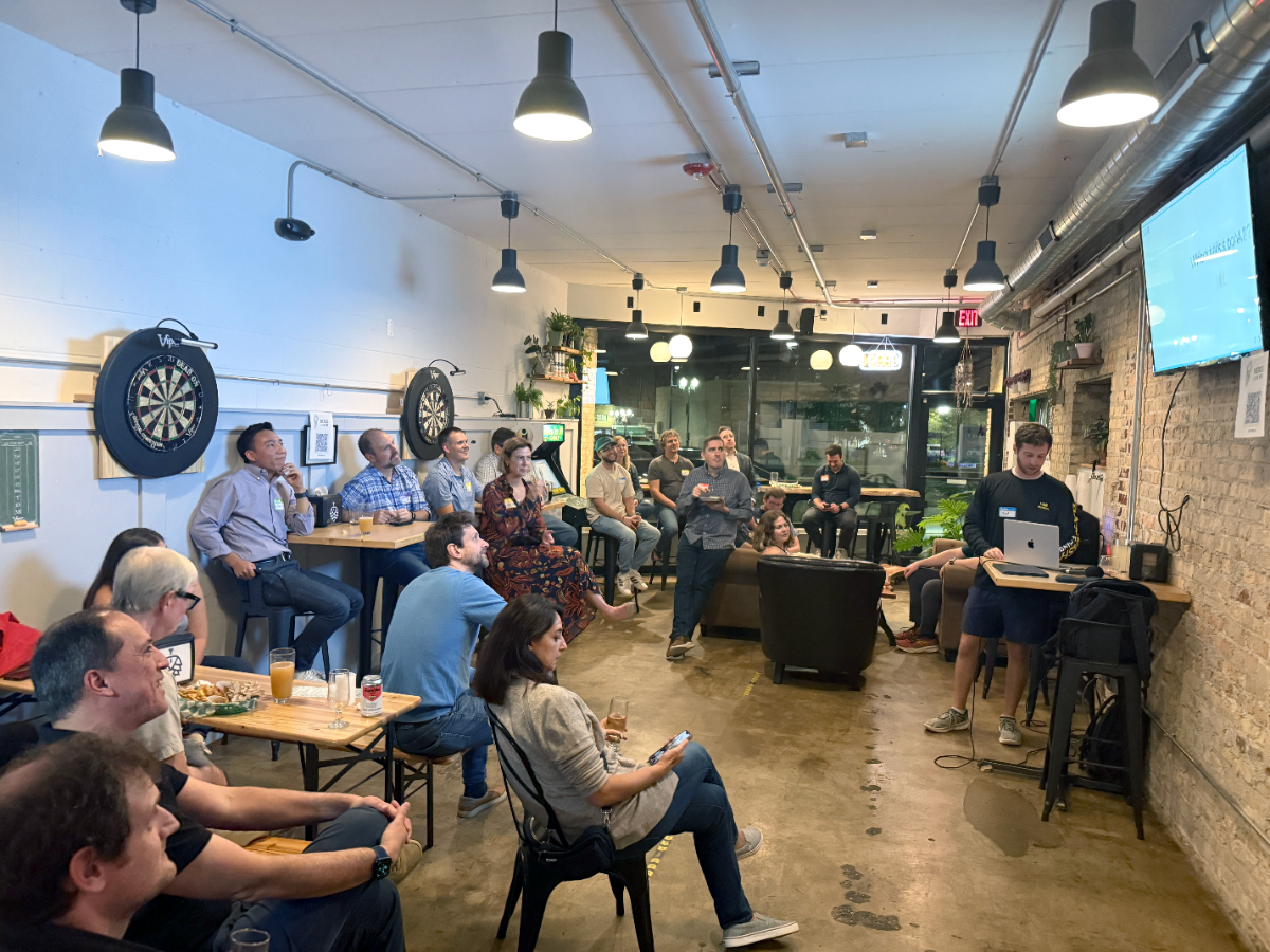 (Photo) A photograph capturing a group of people attending a casual presentation or meeting indoors, set in a venue with exposed brick walls, concrete floors, and industrial lighting. A speaker stands near a large screen and laptop addressing the seated audience. Text: EXIT Group of people attending a presentation or networking event | Indoor venue (likely a bar or event space) with an industrial/loft aesthetic | Dartboards on the wall, exposed brick, concrete floor, overhead pendant lights, and a presenter using a laptop and screen. | Candid, event photography | Colors: #A0785C, #1A1A1A, #E0E0E0 Note: The image is a realistic capture of a real-world scene involving people gathered for an event in an indoor setting, characteristic of a photograph.