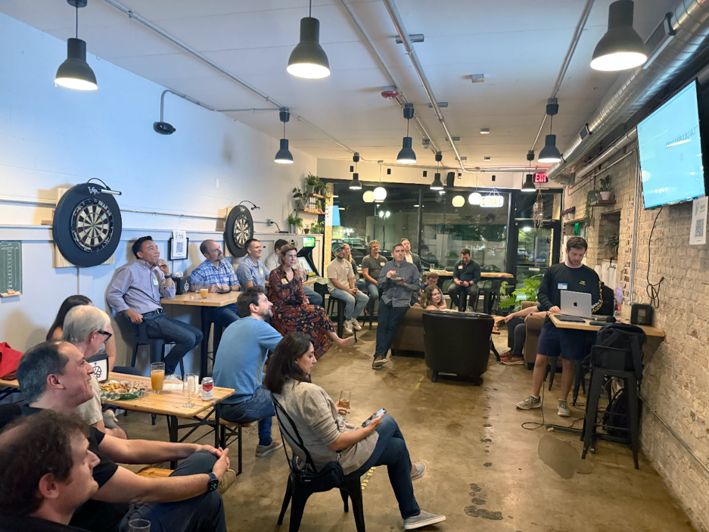 (Photo) A photograph capturing a group of people attending a casual presentation or meeting indoors, set in a venue with exposed brick walls, concrete floors, and industrial lighting. A speaker stands near a large screen and laptop addressing the seated audience. Text: EXIT Group of people attending a presentation or networking event | Indoor venue (likely a bar or event space) with an industrial/loft aesthetic | Dartboards on the wall, exposed brick, concrete floor, overhead pendant lights, and a presenter using a laptop and screen. | Candid, event photography | Colors: #A0785C, #1A1A1A, #E0E0E0 Note: The image is a realistic capture of a real-world scene involving people gathered for an event in an indoor setting, characteristic of a photograph.