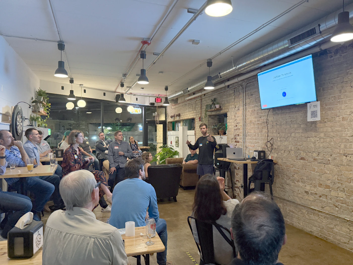 (Photo) The image captures an indoor presentation or meetup event where a speaker addresses an audience gathered in a casual, industrial-style venue with exposed brick walls. Attendees are seated at wooden tables, watching a presentation displayed on a large screen. Text: Portfolio Template EXIT Group of people attending a presentation/lecture | Indoor, casual venue (looks like a bar or co-working space) with exposed brick walls and industrial lighting. | A man speaking and gesturing; a large screen displaying a presentation; numerous people seated; exposed ventilation ducts. | Candid, documentary | Colors: #8B4513, #F5F5DC, #2F4F4F Note: The image is a photograph capturing a real-world event, specifically a speaker giving a presentation to an audience in an indoor setting.