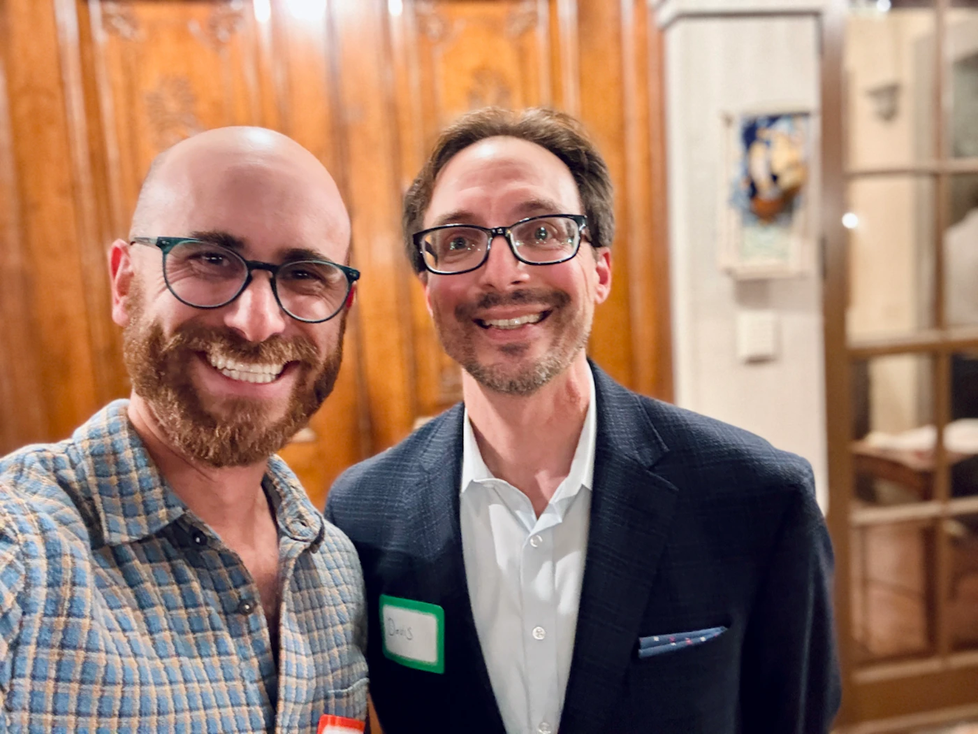 (Photo) A close-up photograph of two men smiling warmly at the camera, likely taken indoors at a social event. They are standing in front of a warm, wood-paneled background and a glass-paned door. Text: Davis Two adult males | Indoor (event space or home interior) | One man wears a plaid shirt and a beard; the other wears a blazer and a name tag reading 'Davis'; warm wood paneling background. | Candid, close-up portrait | Colors: #8B4513, #4682B4, #191970, #F5F5DC Note: The image captures two real individuals posing and smiling at an indoor event, which classifies it as a photograph.
