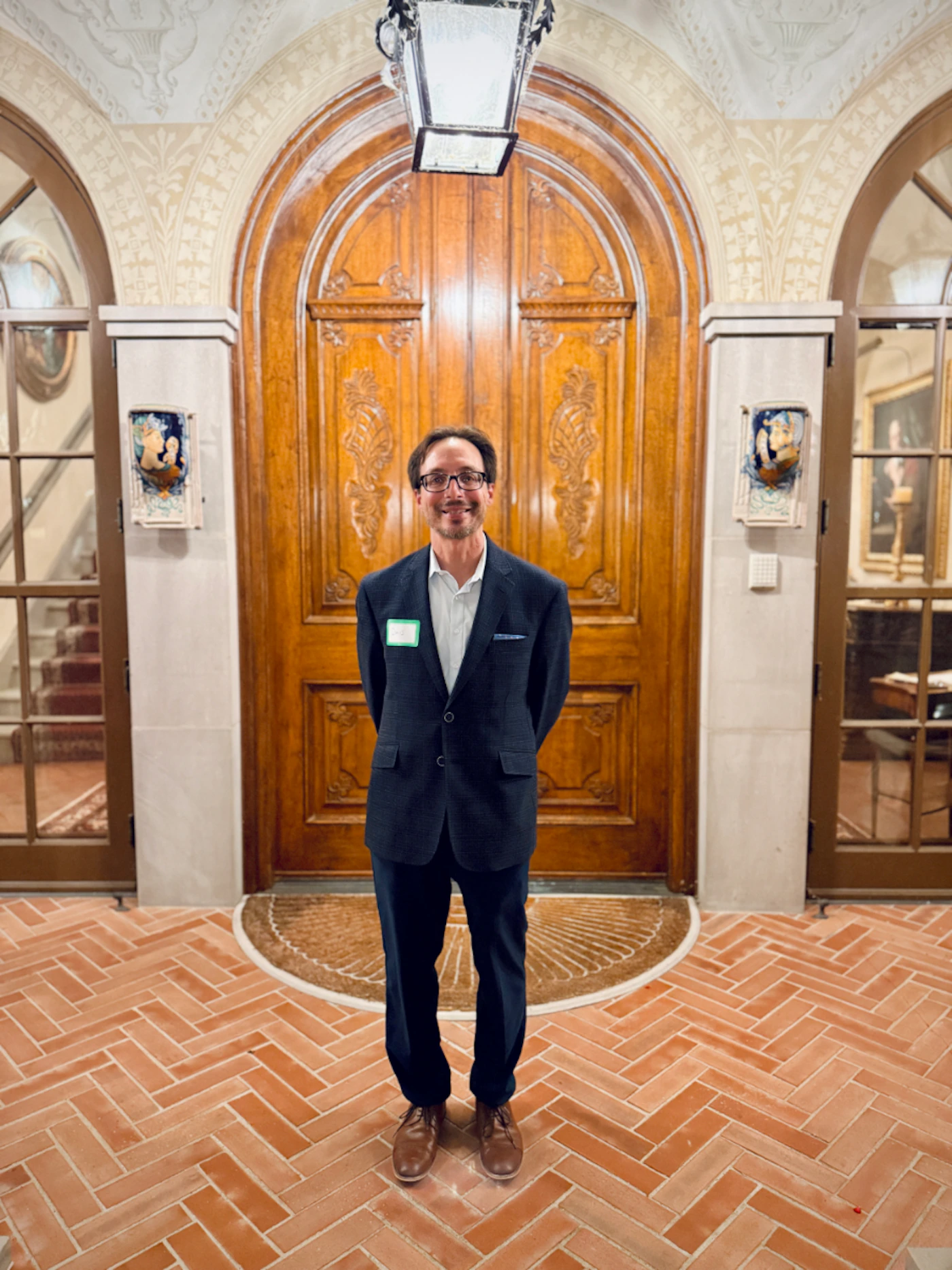 (Photo) A smiling man wearing a dark suit and glasses stands centered in a grand, ornate interior, framed by a large, arched, carved wooden door. A man (full-body portrait) | Indoor, likely a historic building or mansion foyer, characterized by ornate wooden doors and terracotta tile flooring. | Ornate architectural details, strong vertical symmetry, herringbone tile pattern on the floor, and warm ambient lighting. | Full-length portrait, posed, formal setting. | Colors: #A0522D, #36454F, #D2B48C Note: The image is a captured moment of a person standing in an elaborate indoor setting, characteristic of a photograph.