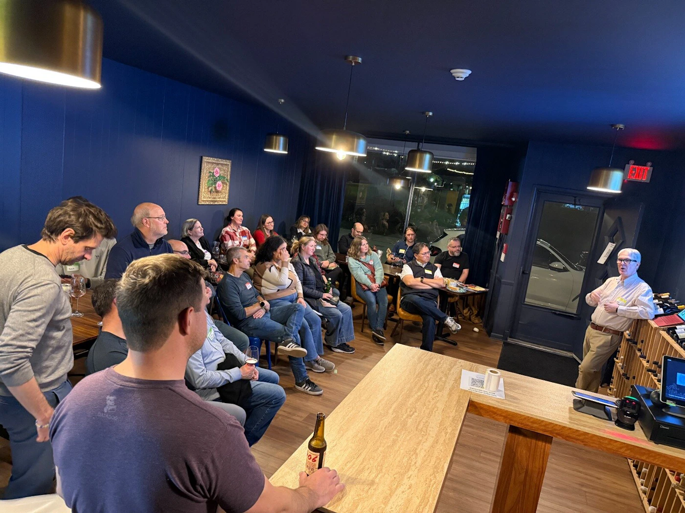 (Photo) A photograph capturing an indoor social or professional gathering where an older man is giving a presentation or talk to an audience seated in a room with dark blue walls and modern lighting. Text: EXIT Group of people attending a presentation/talk | Indoor (event space, likely a bar or wine shop) | Speaker addressing an audience, dark blue walls, brass pendant lights, wine racks visible | Candid, low-light event photography | Colors: #2C3E50, #A0522D, #F5DEB3 Note: The image is a candid photograph documenting a real-world social or professional event taking place indoors, featuring multiple people and a specific setting.