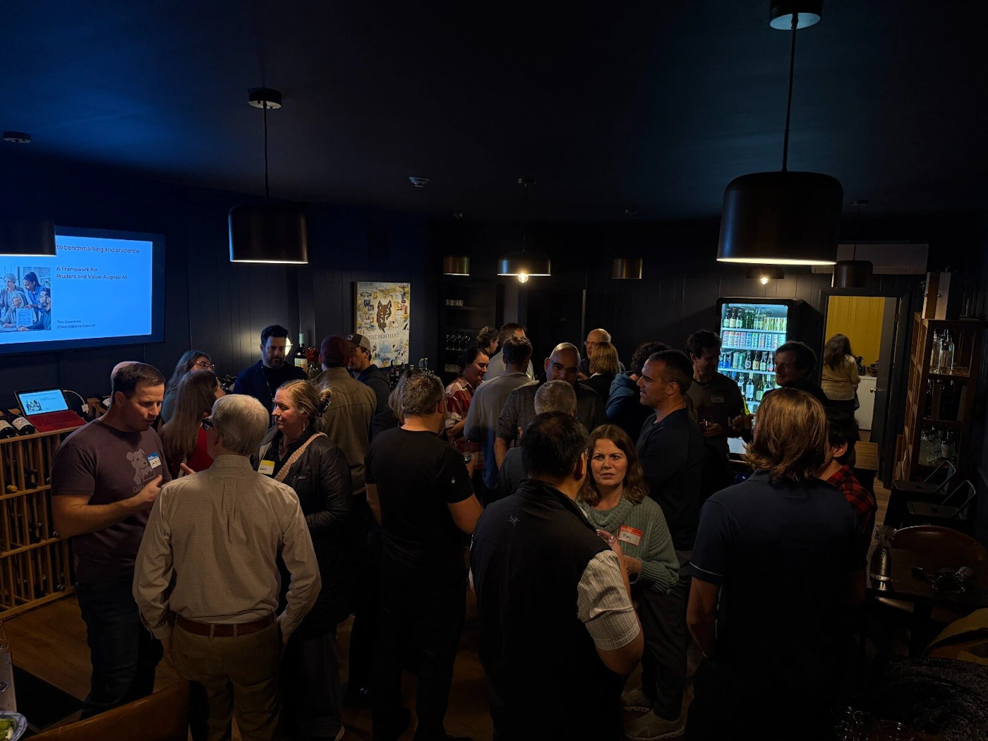 (Photo) A candid photograph captures a large group of people mingling and networking in a dimly lit indoor venue, which appears to be a bar or event space with dark walls, wine racks, and a visible presentation screen on the left. Text: [unreadable] benchmarking and provenance A Framework for Product and Retail Aligned in [unreadable] Group of people, social event, networking | Indoor, dimly lit bar or event space | Large crowd, presentation screen, wine storage, dark interior design | Candid, low-light event photography | Colors: #1E2125, #705C4E, #A0A0A0 Note: The image is a photograph documenting a real-world social event with many people interacting in a specific indoor setting.