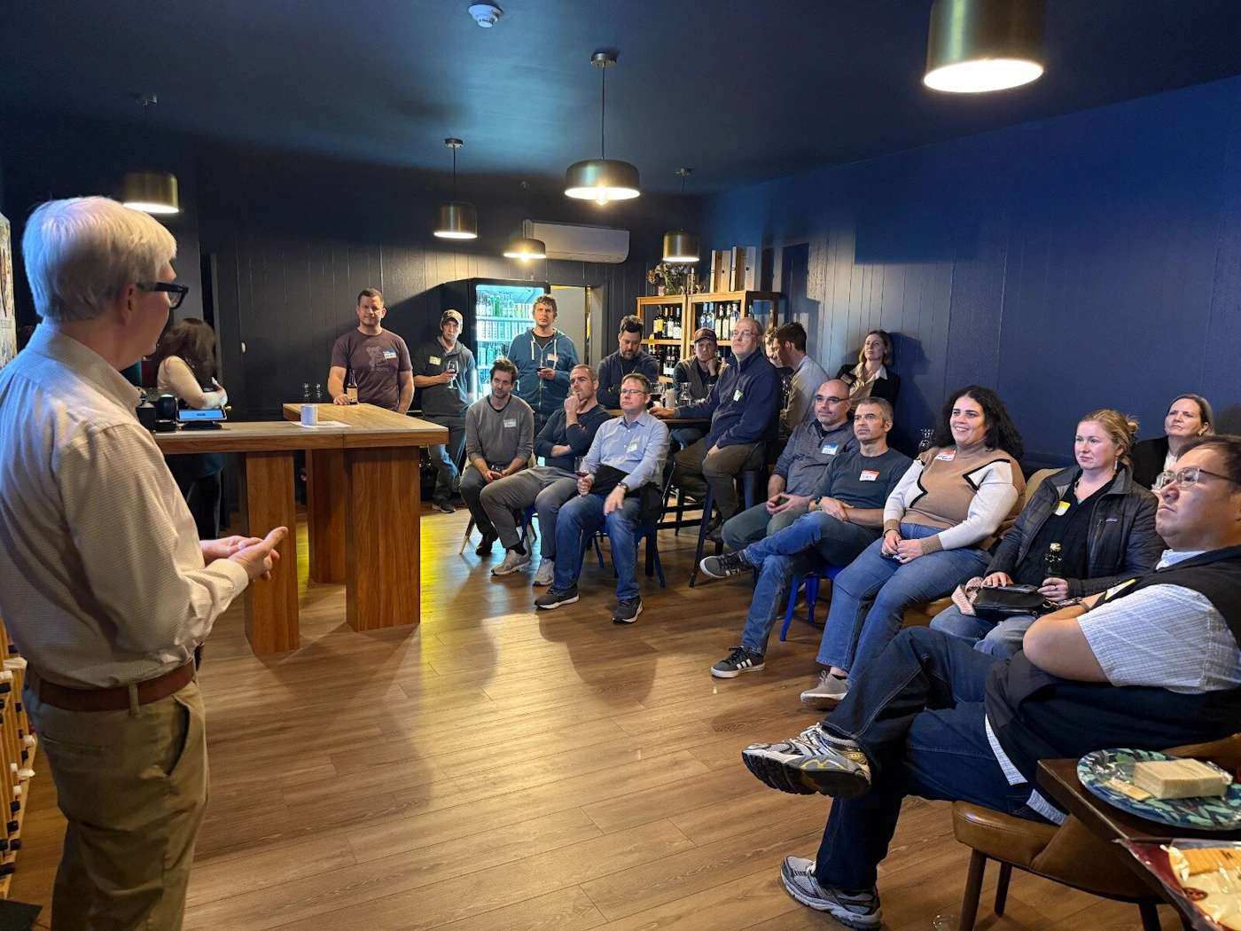 (Photo) A man addresses a seated and standing audience during an indoor gathering or presentation in a dimly lit room featuring dark blue walls and wooden flooring. Group of people attending a presentation or meeting | Indoor (likely a private event space, bar, or meeting room) | Speaker addressing an audience, dark blue walls, wooden table, pendant lighting fixtures. | Candid, event photography | Colors: #2C3A4F, #B89C78, #A3734A, #E0E0E0 Note: The image is a photograph capturing a real-world event involving multiple people in an indoor setting, characteristic of event or candid photography.