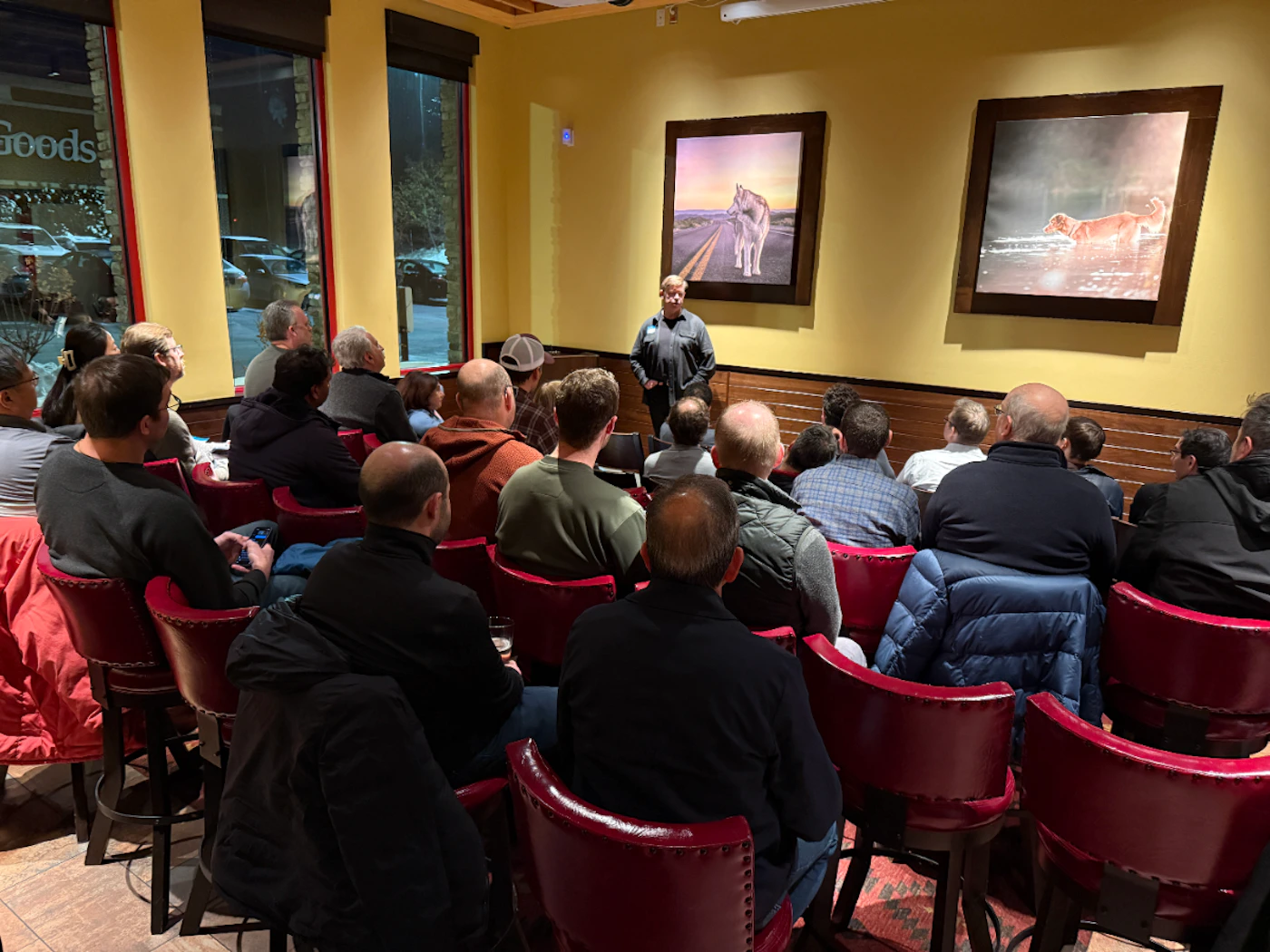 (Photo) A photograph capturing an indoor gathering where an audience, seated in rows of red bar stools, faces a speaker standing against a mustard-yellow wall decorated with two framed pictures of animals. Text: Goods Group of people attending a presentation or meeting | Indoor, likely a private room in a restaurant or bar, at night | Warm, low lighting; red leather bar stools; two prominent framed animal photographs on the wall (a wolf/husky and a dog); snowy outdoor view through large windows. | Candid/event photography | Colors: #A52A2A, #EEDD82, #2F4F4F, #1C1C1C Note: The image is a realistic capture of a moment in time, documenting a group of people attending an event or presentation in an interior setting.