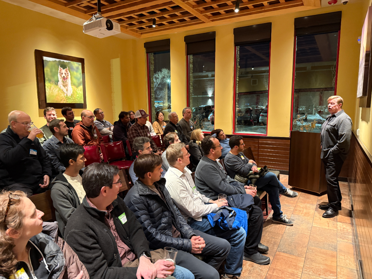 This is a photograph of an indoor gathering where a man is standing and speaking to a seated audience of approximately 30 people. The room has mustard-yellow walls, a wooden lattice ceiling, and three tall windows showing a dark outdoor scene.
