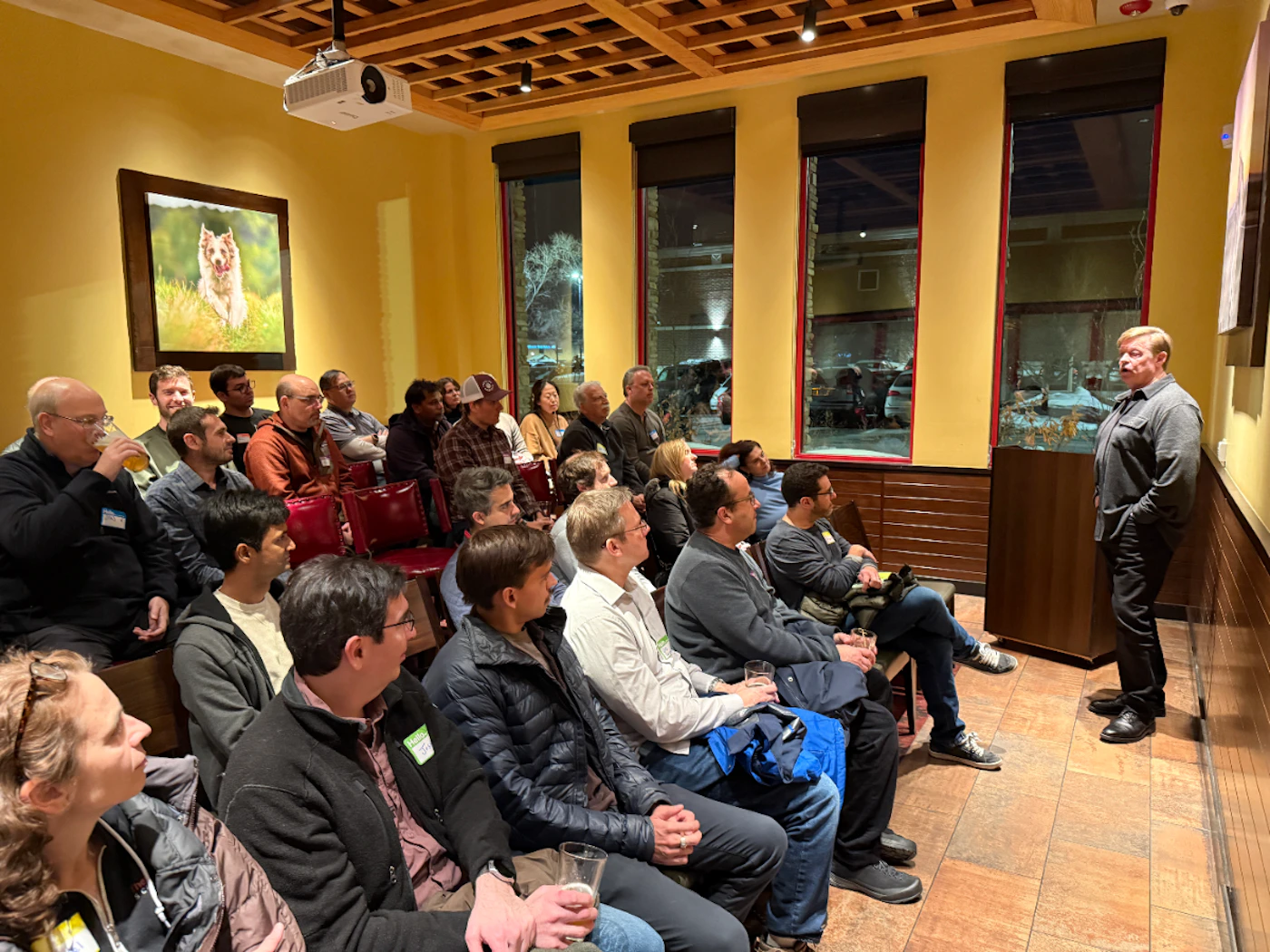 (Photo) This is a photograph of an indoor gathering where a man is standing and speaking to a seated audience of approximately 30 people. The room has mustard-yellow walls, a wooden lattice ceiling, and three tall windows showing a dark outdoor scene. A speaker addressing a large audience during an indoor event or meeting. | Indoor meeting room or restaurant space, during the evening or night. | A projector is mounted on the ceiling, a large framed photo of a dog is on the wall, and many attendees are holding drinks. | Candid, event documentation, low-light setting. | Colors: #d6b34a, #4d4e4e, #7f3b3b, #2c2c2c Note: The image is a realistic capture of a real-world scene involving people attending an event or presentation, classifying it as a photograph.