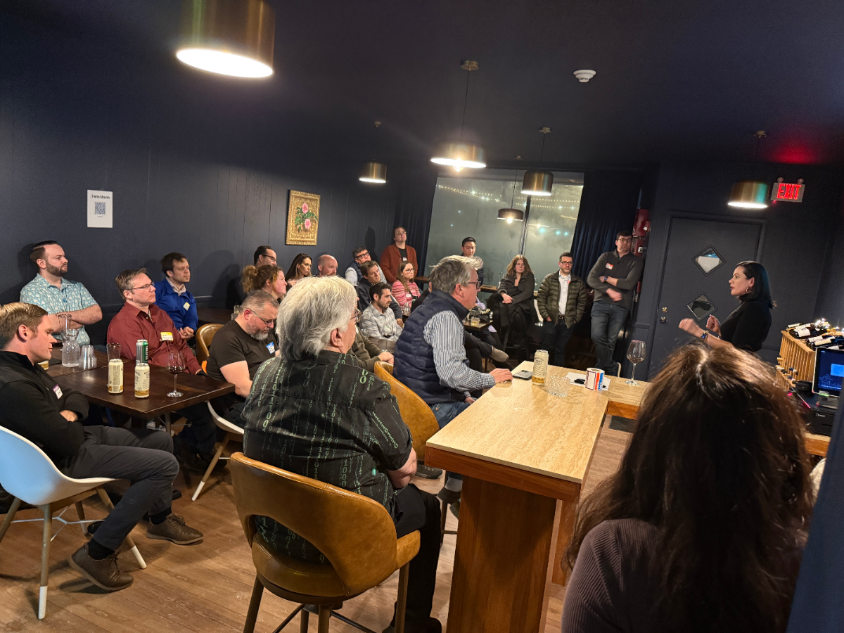 (Photo) A photograph capturing an indoor event or presentation held in a dimly lit venue with dark blue walls, where a large group of people is seated and standing, listening attentively to a woman who is speaking near a wooden counter. Text: EXIT Group of people attending an event or presentation | Indoor, dark event space or bar | Dim lighting, multiple people seated at tables with drinks, one speaker addressing the group. | Candid, event photography | Colors: #1C2533, #D2B48C, #A1754B, #FFFFFF Note: This image is a direct capture of a real-world scene—an indoor gathering—making it a photograph.