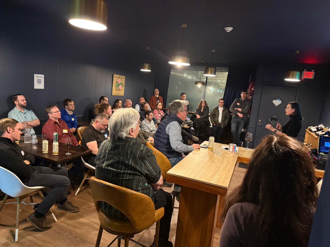 (Photo) A photograph capturing an indoor event or presentation held in a dimly lit venue with dark blue walls, where a large group of people is seated and standing, listening attentively to a woman who is speaking near a wooden counter. Text: EXIT Group of people attending an event or presentation | Indoor, dark event space or bar | Dim lighting, multiple people seated at tables with drinks, one speaker addressing the group. | Candid, event photography | Colors: #1C2533, #D2B48C, #A1754B, #FFFFFF Note: This image is a direct capture of a real-world scene—an indoor gathering—making it a photograph.