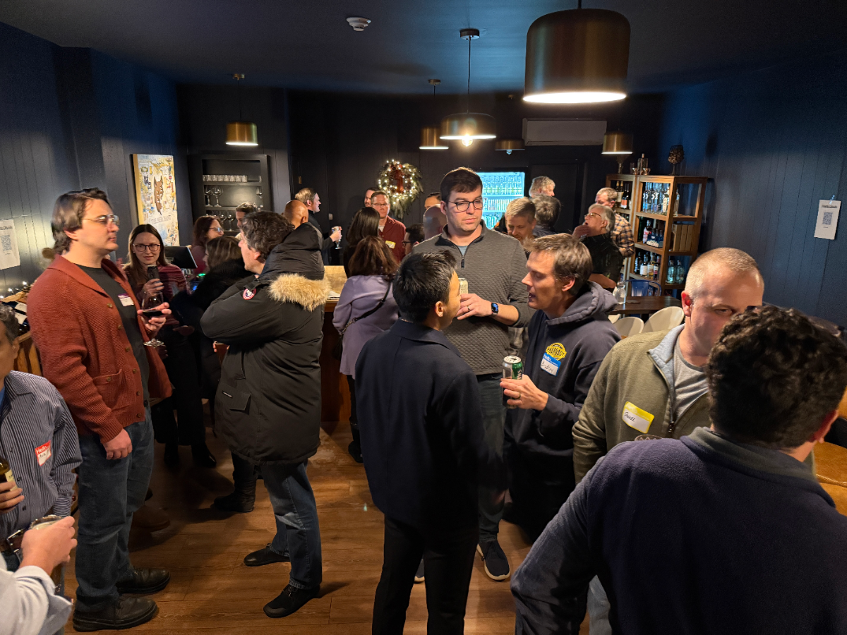 A photo capturing a social gathering or networking event indoors, where a large group of people are standing, talking, and holding drinks in a dimly lit room with dark blue walls and modern light fixtures.. Text: [unreadable]