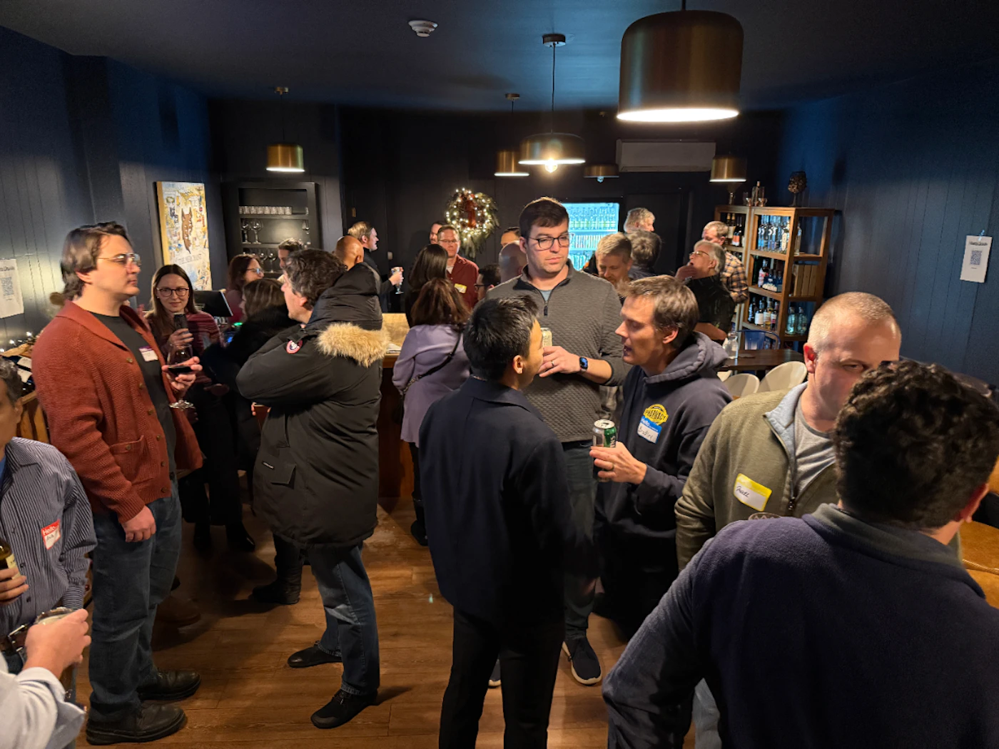 (Photo) A photo capturing a social gathering or networking event indoors, where a large group of people are standing, talking, and holding drinks in a dimly lit room with dark blue walls and modern light fixtures. Text: [unreadable] Group of people socializing | Indoor, dark event space or private room | Large crowd, name tags visible on clothing, dim lighting, holiday wreath in background. | Candid, low-light event photography | Colors: #2D3748, #8B4513, #A52A2A, #D3D3D3 Note: This is a candid photograph documenting an indoor social event where people are mingling and interacting, characteristic of event photography.