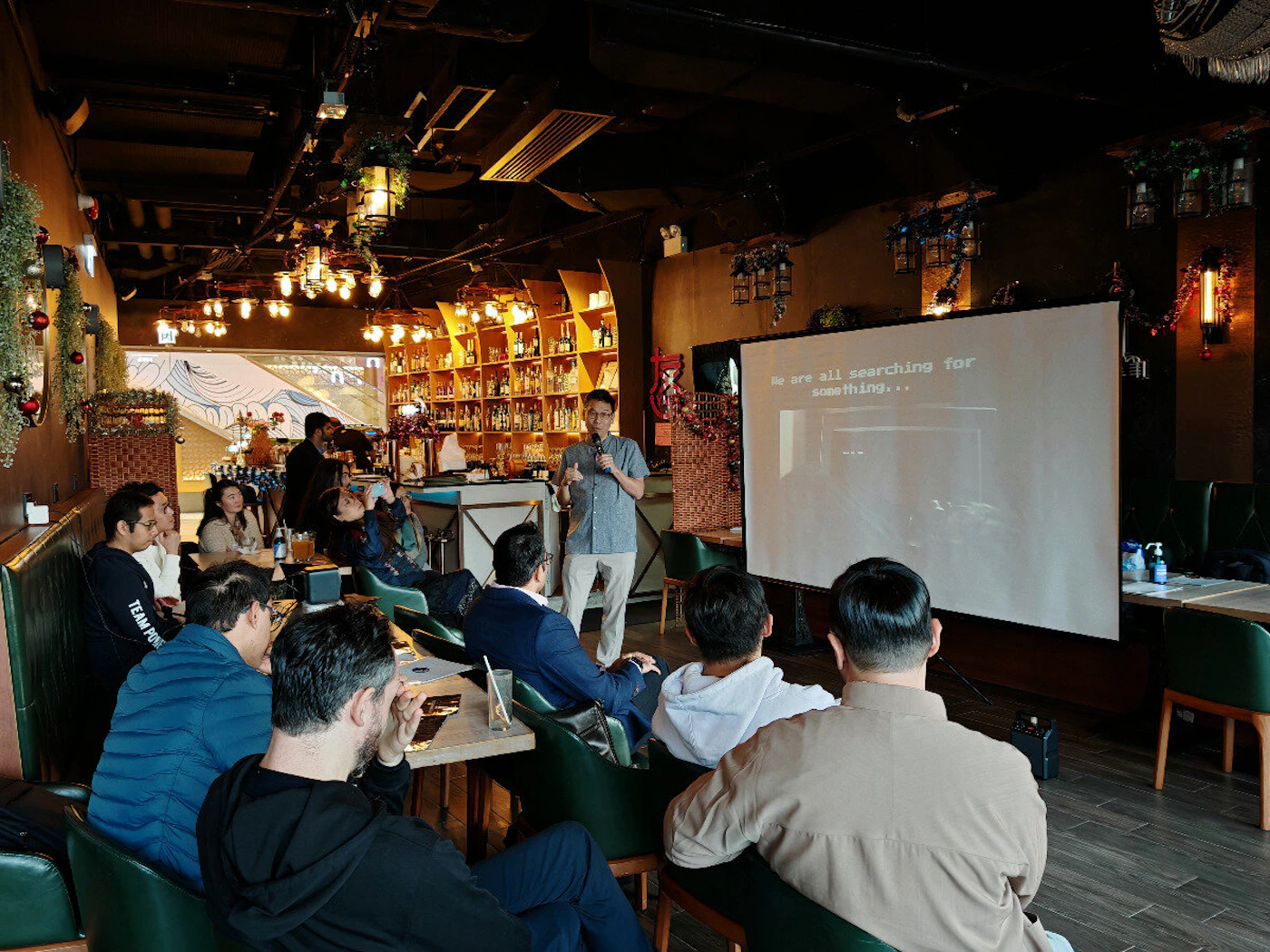 (Photo) A man is giving a presentation to a group of people in a bar or restaurant.  He is standing in front of a projector screen displaying text. Text: We are all searching for something... people | indoor | presentation, projector screen, audience | candid Note: The image is a real-life captured scene of a presentation, depicting people and a setting. This makes it a photograph.