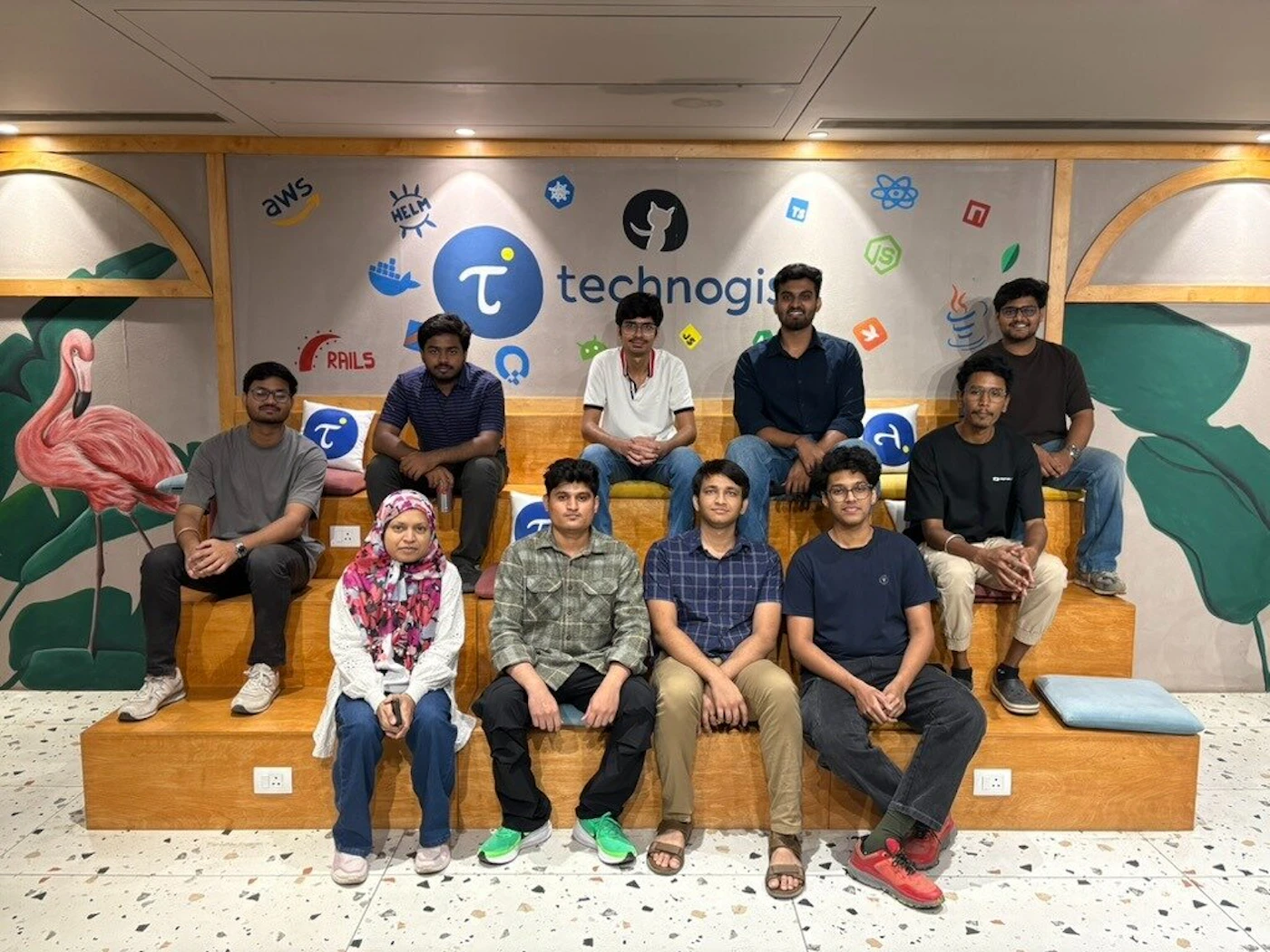 (Photo) A group of ten young people, nine men and one woman, are posing for a group photo while sitting on a tiered wooden bench in an indoor office setting. Text: aws, HELM, technogis, RAILS, JS, TS, Java group of people | indoor office space | tiered wooden seating, wall decorated with various tech logos and the company name 'technogis' | group portrait | Colors: #C08E5F, #3A5A97, #FFFFFF, #000000 Note: The image is a real-life photograph of a group of people in a professional or educational environment.