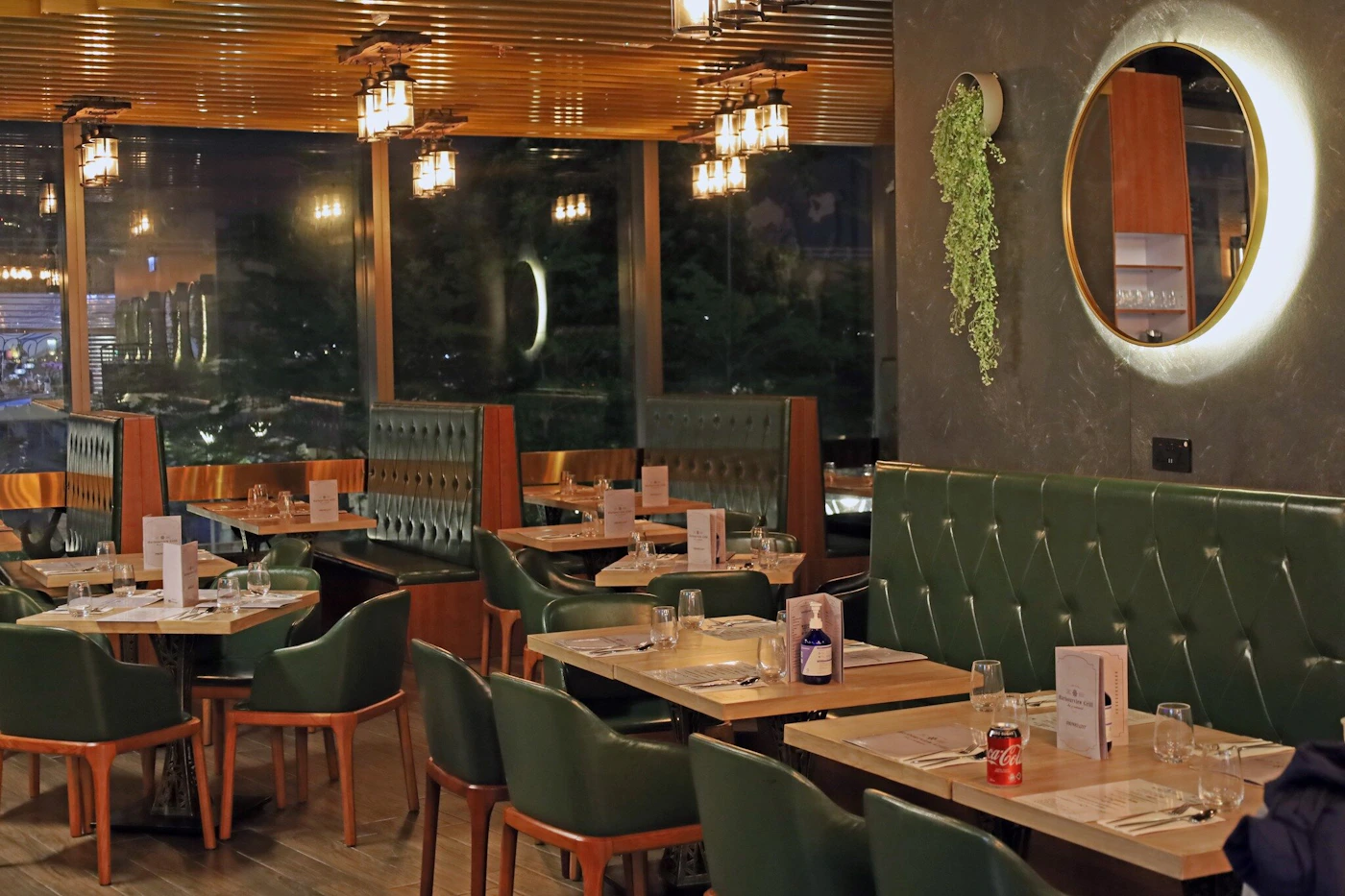 (Photo) A photo of a restaurant interior with green tufted booths, wooden tables set for dining, and a view of a cityscape through large windows at night. restaurant interior | indoor | tufted booths, city view, night scene | interior photography Note: The image is a realistic depiction of a physical space, captured through photography. It showcases the restaurant's ambiance and interior design.