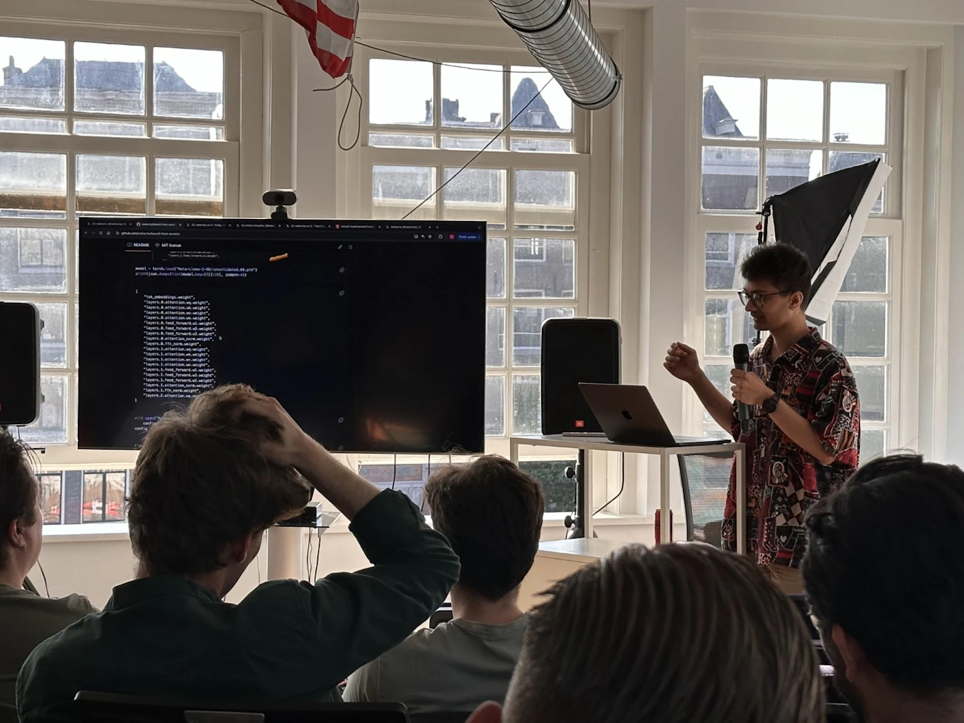 (Photo) A person is presenting to a small group of people, with a large screen displaying code in the background. people | indoor | presentation, audience, code on screen | candid Note: The image is a real-life captured scene of a presentation, depicting people and a setting, characteristic of a photograph.