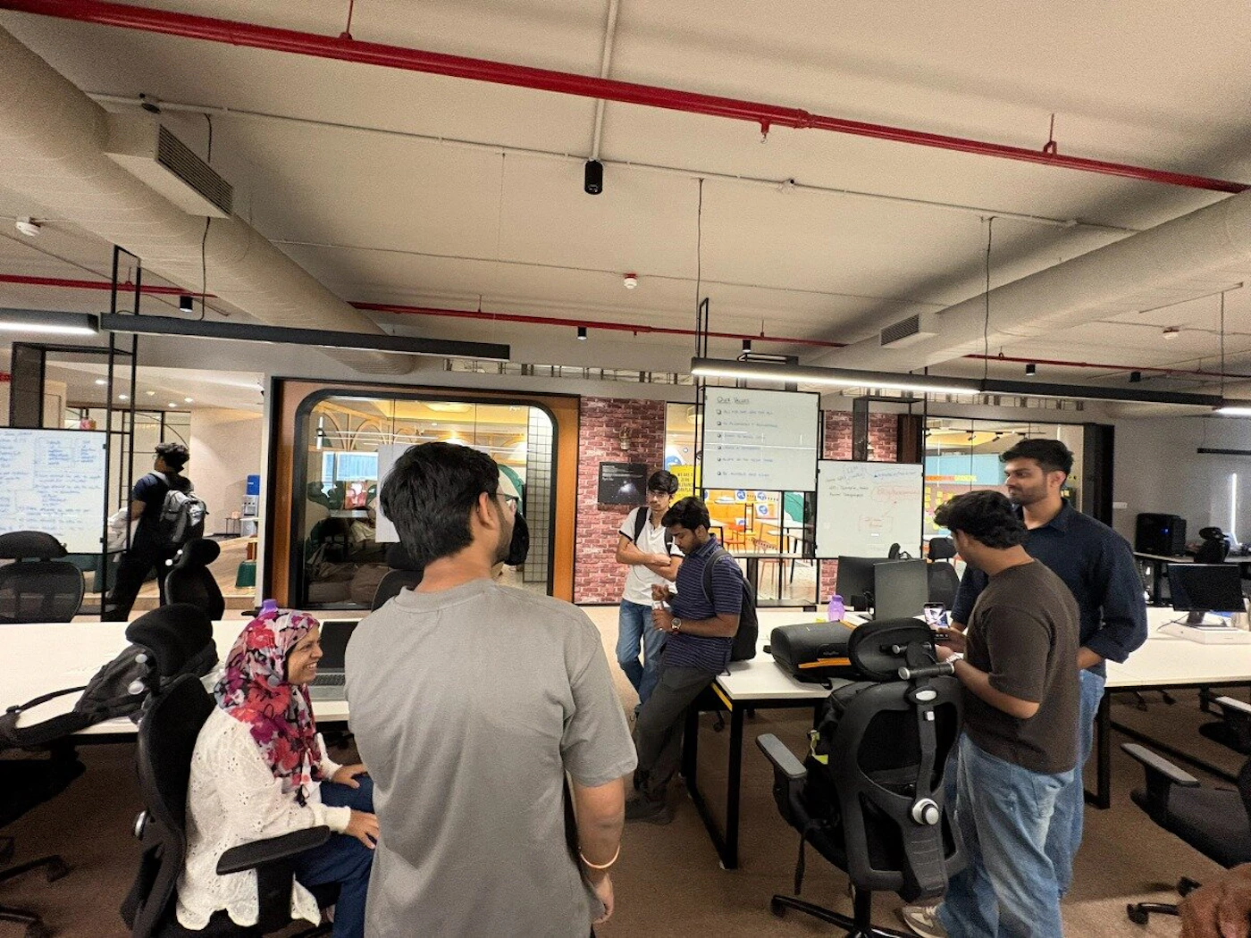 (Photo) A group of people are gathered in a modern office or coworking space, with some standing and talking while one woman is seated. The room features whiteboards, workstations, and prominent red pipes on the ceiling. Text: Our Team The Founders of [unreadable] Head of [unreadable] Head of [unreadable] Head of [unreadable] Head of [unreadable] Head of [unreadable] Head of [unreadable] People in an office | indoor office/coworking space | Multiple people, whiteboards with text, modern industrial ceiling with red pipes, office chairs and desks. | candid | Colors: #FFFFFF, #808080, #000000, #FF0000 Note: This is a real-life photograph capturing a candid moment of people in an indoor office environment.