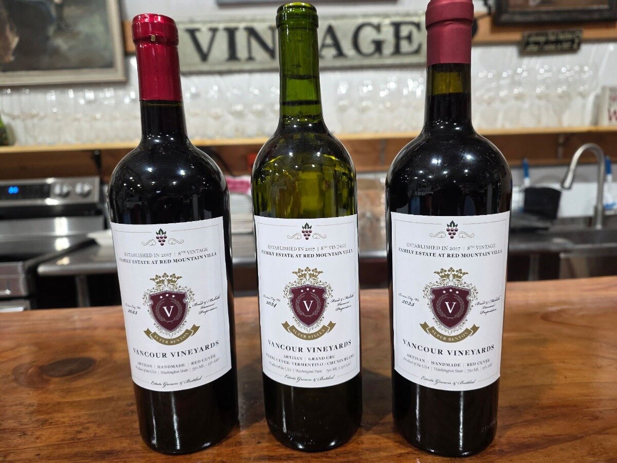 A close-up photograph shows three bottles of Vancour Vineyards wine standing on a wooden bar counter in an indoor setting, likely a tasting room. The bottles feature white labels with a prominent red and gold crest.. Text: VINTAGE, ESTABLISHED IN 2017 | 8TH VINTAGE, FAMILY ESTATE AT RED MOUNTAIN VILLA, 2023, SILVER BENTON, VANCÕUR VINEYARDS, ARTISAN HANDMADE RED CUVÉE, Estate Grown & Bottled, Washington State, 750 ML, GRAND CRU, CABERNET SAUVIGNON