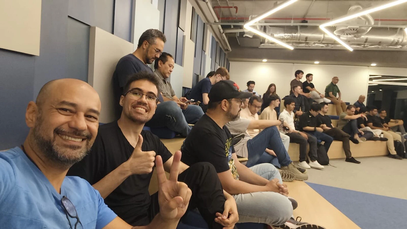 (Photo) A candid photo taken from the front row of a tiered seating area, showing a large group of people attending an indoor event or presentation. Two men in the immediate foreground are smiling and posing for the camera with a thumbs up and a peace sign. Group of people attending an event/presentation | Indoor, modern office/auditorium space with tiered seating | Large group, selfie perspective, tiered seating, modern architecture | Candid/Selfie | Colors: #4173A9, #1A1A1A, #D5C1A2, #FFFFFF Note: The image is a captured moment of real people in a real-world setting (an indoor event space), characteristic of a photograph.