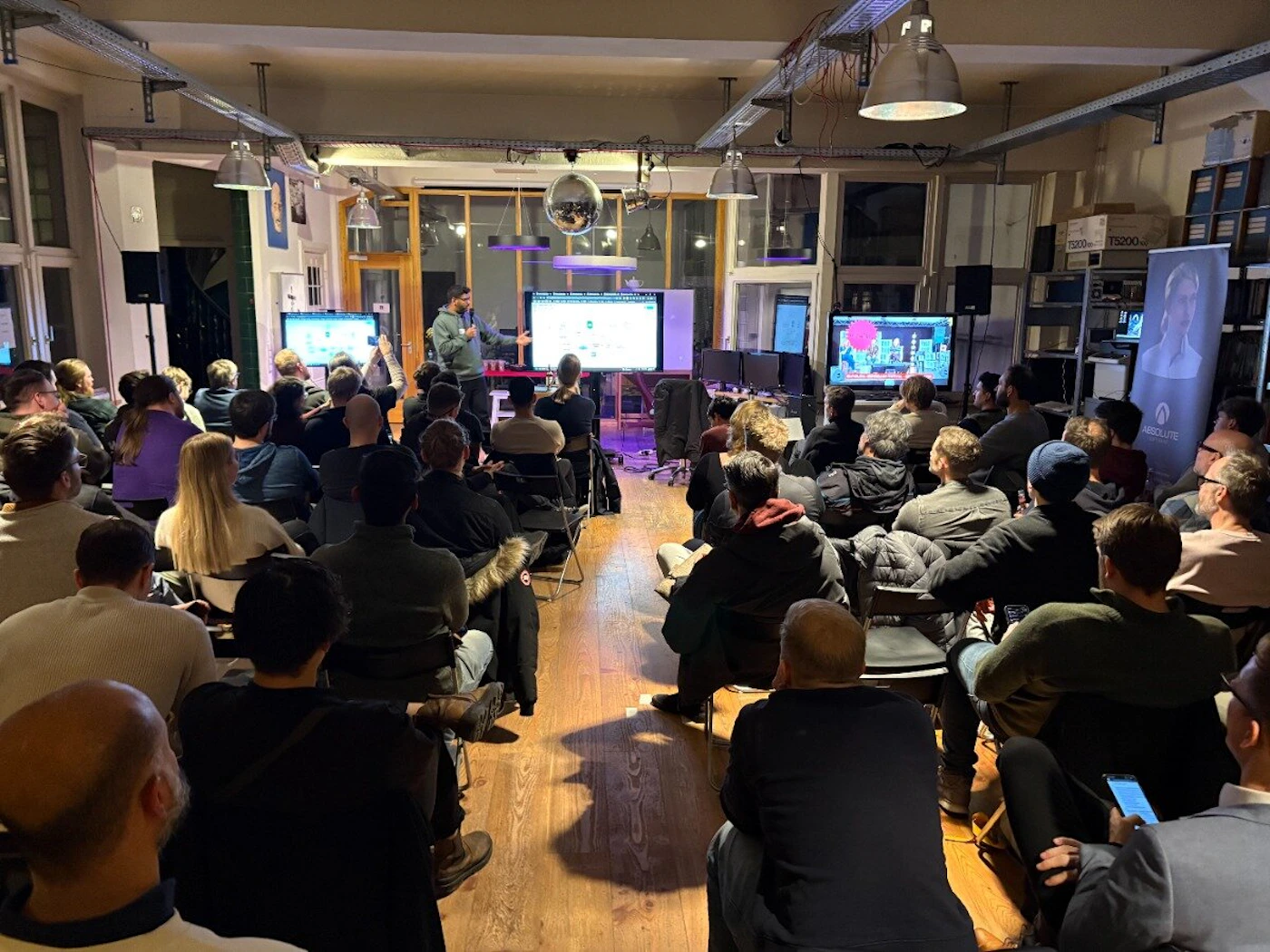 (Photo) A wide shot photograph captures an indoor event where a group of people are seated, facing a speaker who is presenting in front of two large illuminated screens. The room has an industrial aesthetic with exposed conduits, a disco ball, and warm lighting. Text: ABSOLUTE people attending an event or presentation | indoor, likely an event space or large room with an industrial design | multiple people in an audience, a speaker, two large presentation screens, a disco ball, and visible lighting fixtures | candid, event photography, wide shot | Colors: #2C2C2C, #B07B4F, #DCDCDC, #9933FF Note: The image is a direct capture of a real-world scene, showing people and an indoor environment, consistent with a photographic representation.
