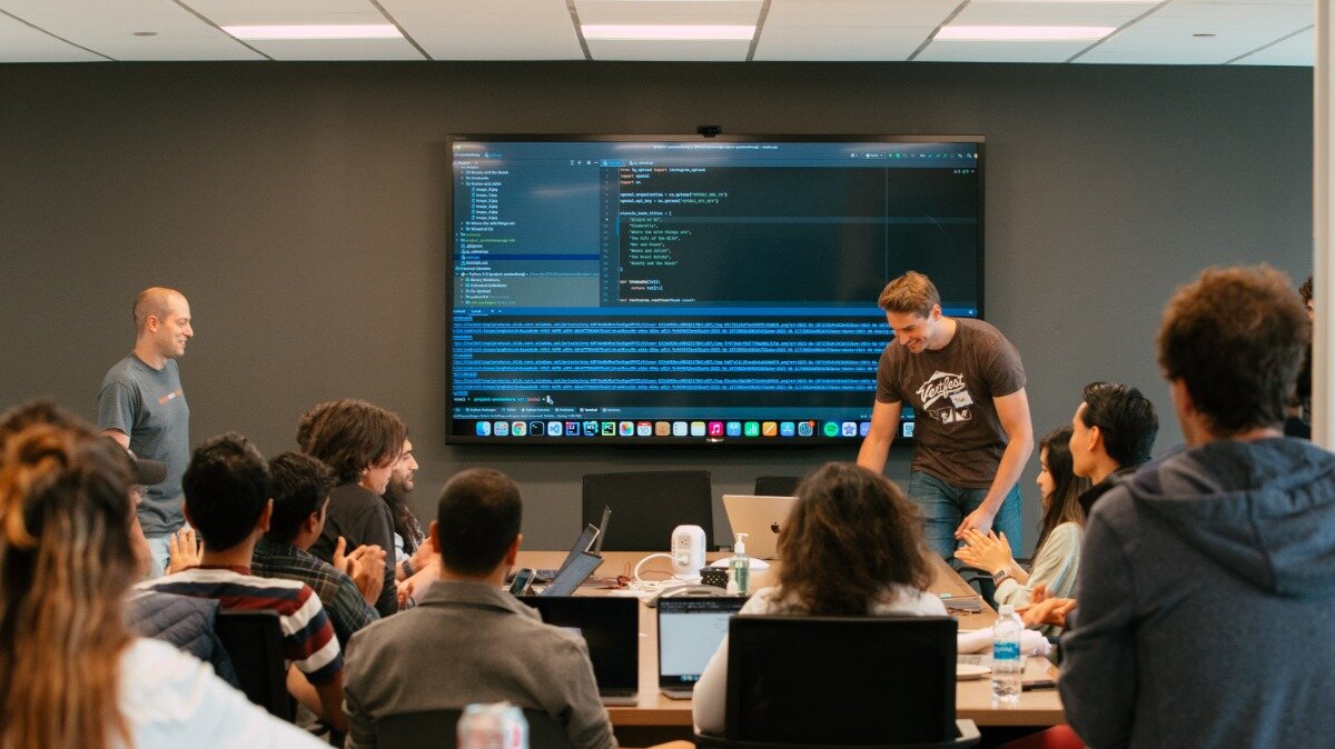 (Photo) A group of people are gathered in a modern conference room, attentively watching a presentation on a large screen that displays lines of code. A man in a brown t-shirt is standing near the screen, seemingly explaining something, while another man in a gray t-shirt stands to the left, observing. Text: [unreadable code and application names on screen], [unreadable text on t-shirts] people, meeting, presentation, technology | indoor, conference room, office | multiple people, large screen displaying code, laptops on a table, modern interior | candid, documentary | Colors: #4A4A4A, #8B572A, #1E1E1E, #D4D4D4, #9CDCFE Note: The image is a realistic depiction of a scene captured by a camera, showing people and an environment in a naturalistic style, fitting the definition of a photograph.