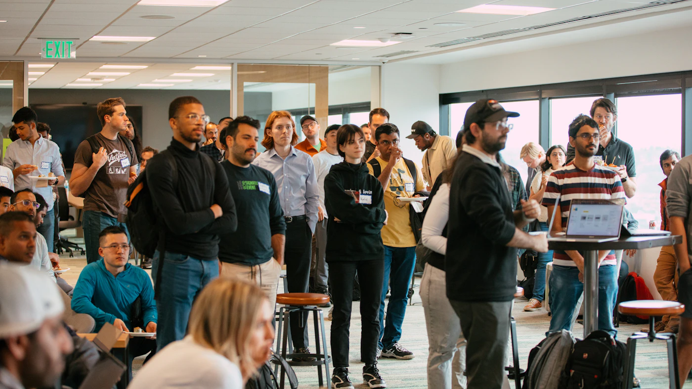 (Photo) A photograph of a group of people gathered in an office space, seemingly attending a presentation or meeting. Text: EXIT people | indoor | group of people, office setting, presentation/meeting | candid Note: The image is a real-life captured scene of people in an office environment, indicating a photograph.  It depicts a spontaneous moment rather than a posed shot.