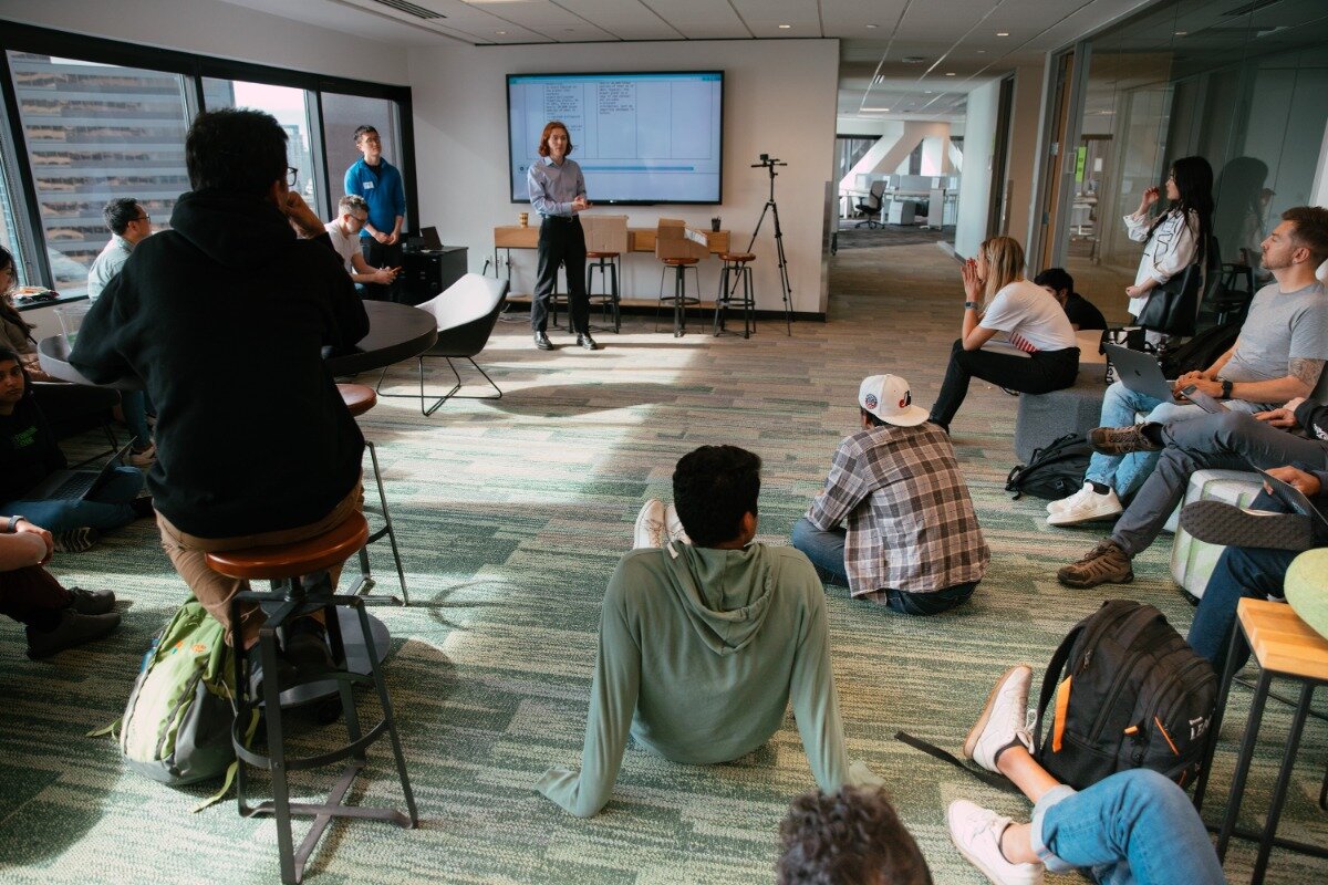 (Photo) A group of people are gathered in a modern, open-plan office space, attending what appears to be a presentation or meeting. A person stands at the front addressing the group, with a large screen displaying information behind them, while others are seated casually on the floor, stools, and low furniture. A group of people attending a presentation or meeting | Indoor, modern office or co-working space with large windows | Multiple people, a presenter, a large display screen, casual seating arrangements including floor seating and stools, and an open-plan office layout. | Candid, documentary, or event photography | Colors: #87B19F, #4F5F5C, #D0D0D0, #3A3A3A, #7C573A Note: The image is a realistic capture of a scene with people and an environment, indicating it is a photograph rather than a graphic design, illustration, or text-based image.