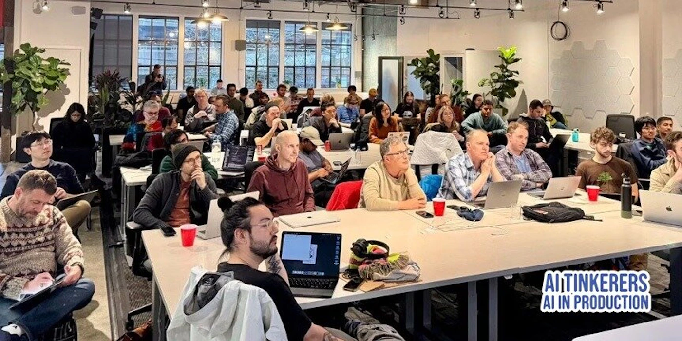 (Photo) A photo of a large group of people sitting at tables in a workshop or conference setting, seemingly focused on laptops. Text: AItinkerers AI IN PRODUCTION people | indoor | large group, laptops, tables, workshop/conference setting | candid Note: The image is a real-life captured scene of people in a room, indicative of a photograph. The presence of laptops and the arrangement suggest a workshop or conference.