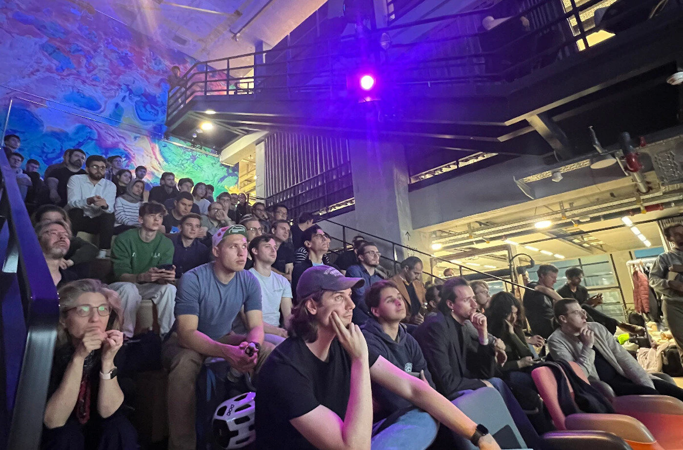 (Photo) A photo of a large group of people sitting on bleachers in an indoor space, appearing to watch a presentation or performance. people | indoor | large group, bleachers, presentation setting | candid Note: The image is a real-life captured image of people in a specific setting, making it a photograph.  It depicts a scene rather than a logo, banner, illustration, or handwriting.