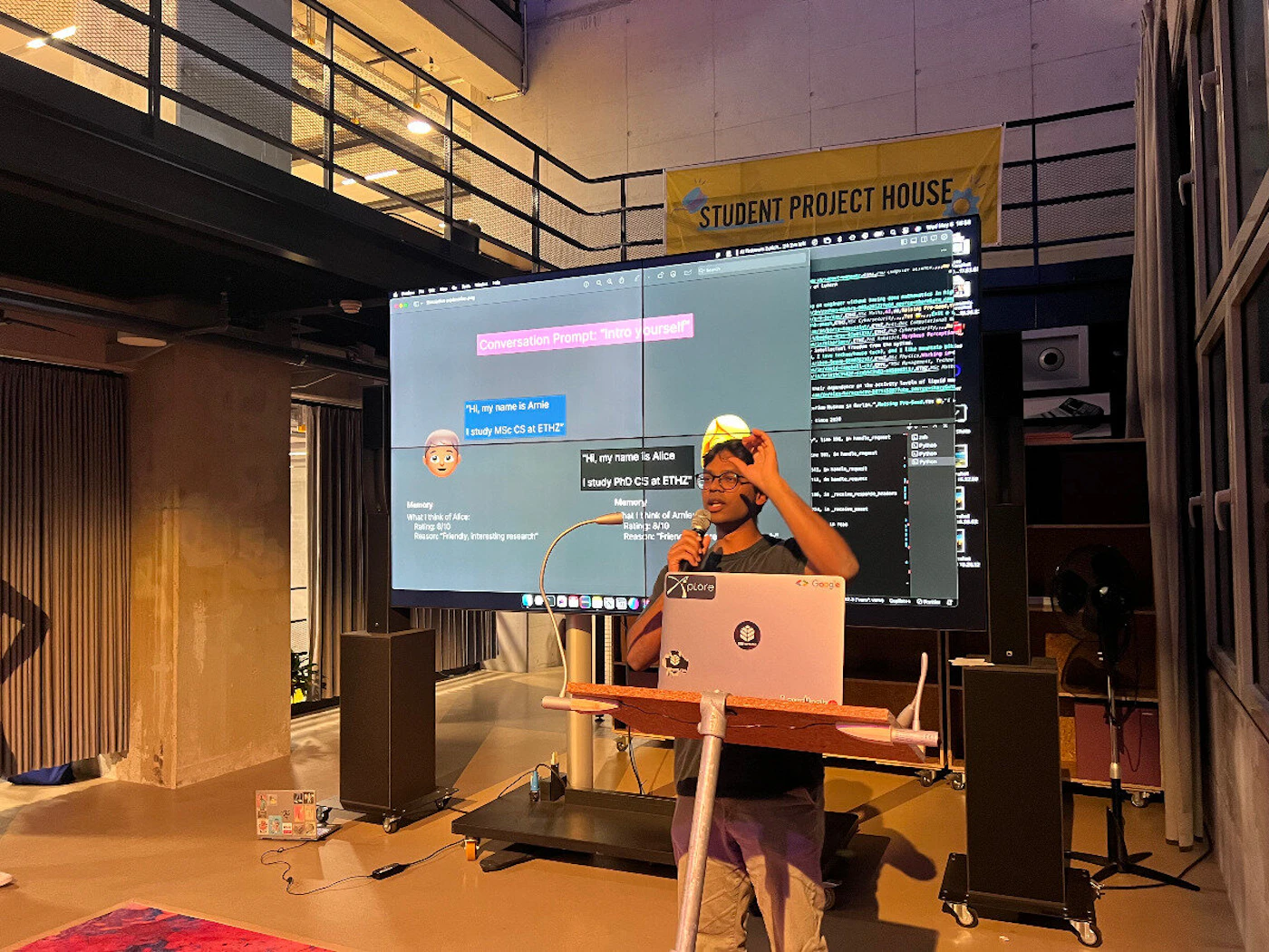 (Photo) A person is giving a presentation about a conversational AI project in a room with a large screen displaying code and conversation prompts. Text: Conversation Prompt: ... "Hi, my name is Alice I study MSc CS at ETH" "Hi, my name is Bob I study PhD CS at ETHZ" STUDENT PROJECT HOUSE person | indoor | presentation, screen with code and conversation, audience | candid Note: The image is a real-life captured scene of a presentation, depicting a person, a screen with content, and the surrounding environment. This makes it a photograph.