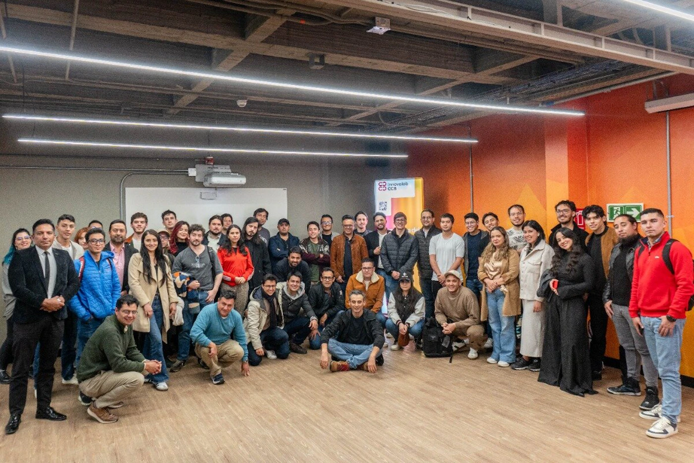 (Photo) A large group of approximately 40 people, consisting of men and women, pose closely together for a group photo indoors in what appears to be a modern event or classroom space. Text: innovalab OCR Group portrait/People | Indoor (event space/classroom) | Large group, mixed genders, modern interior design with orange accent wall and strip lighting. | Group portrait, posed | Colors: #8D6E63, #FF5722, #333333, #CCCCCC Note: The image is a photographic capture of a real-world scene, specifically documenting a large group of people gathered indoors for an event.