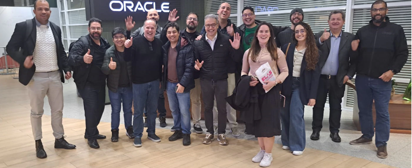(Photo) A group of approximately 15 people are posing for a photo indoors, likely in an office or event space, with some smiling, waving, or giving thumbs up. A large 'ORACLE' sign is visible in the background. Text: ORACLE ELAS group of people | indoor, possibly an office building or event hall | multiple people, diverse ages and genders, some gesturing (waving, thumbs up), large company sign in background | group portrait, candid | Colors: #1E1E1E, #F0F0F0, #4A6E9B, #D4C3A9, #C79F7F Note: The image captures a moment with a group of individuals in a real-world setting, characteristic of a photograph. It is not an illustration, logo, banner, or handwriting.