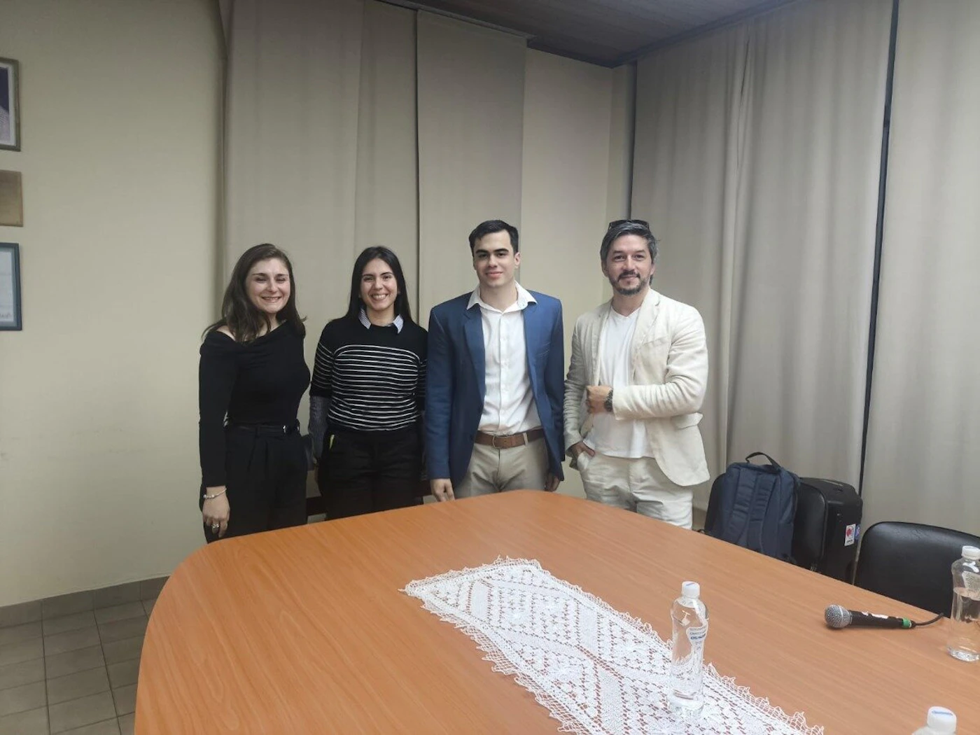 (Photo) A group of four people, two women and two men, are standing behind a large wooden table in an indoor setting, likely a meeting or conference room. The table is adorned with a white lace runner, water bottles, and a microphone. Four individuals (two women, two men) and a meeting room setup. | Indoor, likely a conference room or classroom. | A group of people posing for a photo, a large oval wooden table, a white lace table runner, water bottles, a microphone, and a backpack. | Group portrait/candid. | Colors: #8B4513, #E0DEDC, #000000, #3A5F8B, #F0EAD6 Note: The image is a realistic capture of a scene featuring people and objects in an indoor environment, typical of a photograph rather than an illustration, logo, or banner.