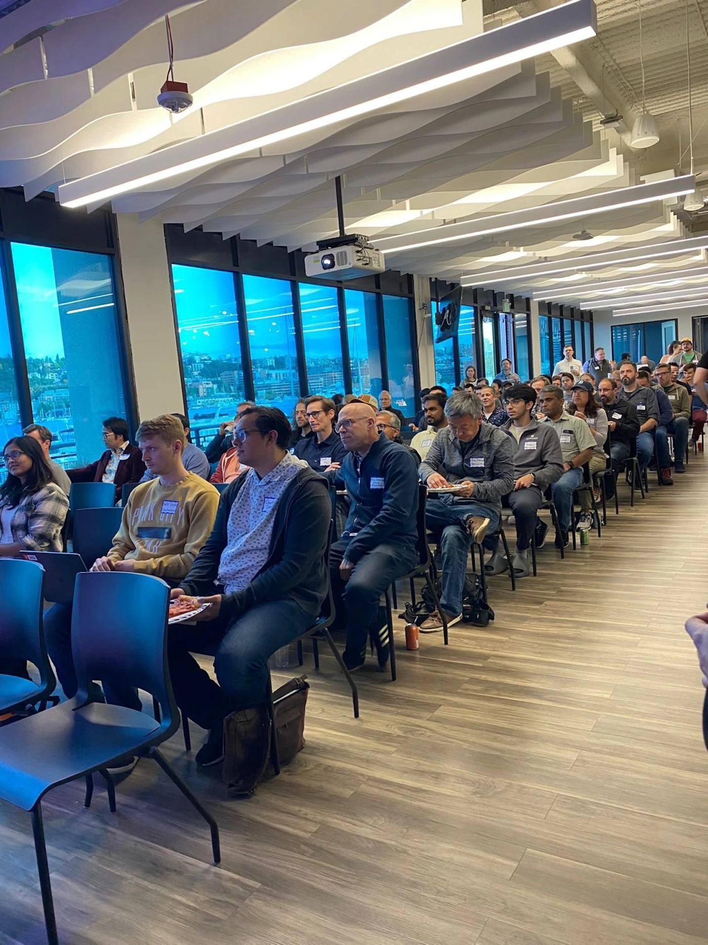 (Photo) A photo of a large group of people sitting in rows of chairs in a modern office space, seemingly attending a presentation or meeting. people | indoor | large group, presentation setting | candid Note: The image is a real-life captured scene of people in an indoor setting, indicative of a photograph. It's not a drawing, logo, banner, or handwriting.