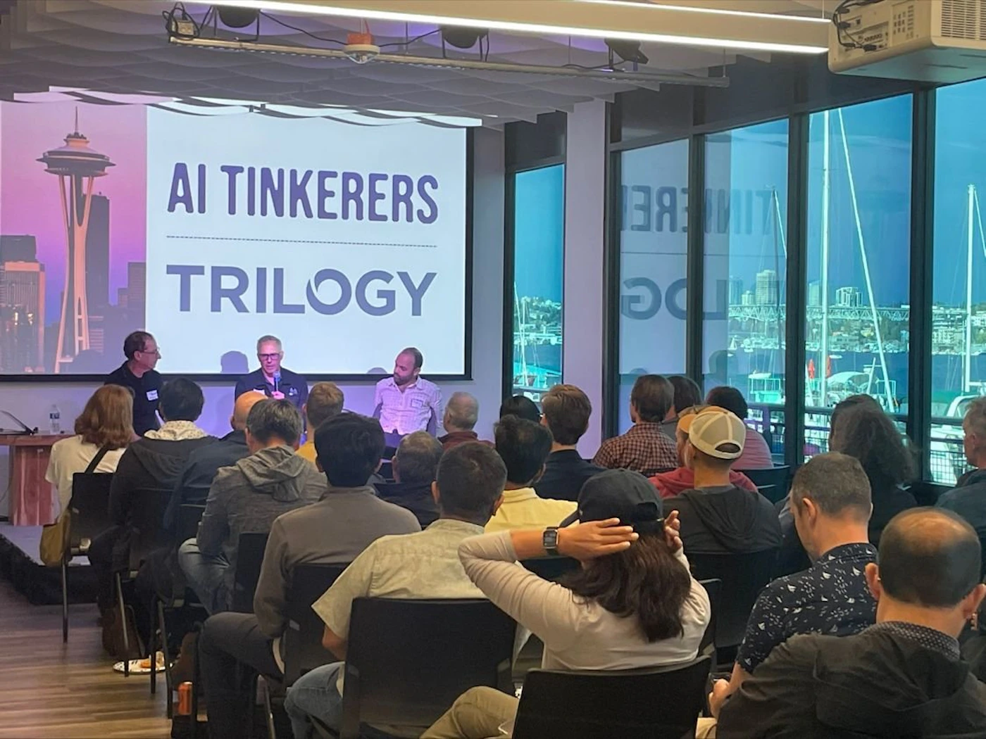 (Photo) A photo of an audience watching a presentation by three people on a stage with a projected image featuring the Space Needle and the text 'AI Tinkers' and 'Trilogy'. Text: AI TINKERERS TRILOGY people | indoor | presentation, audience, stage | candid Note: The image is a real-life captured scene of an event, depicting people and a presentation setting, making it a photograph.