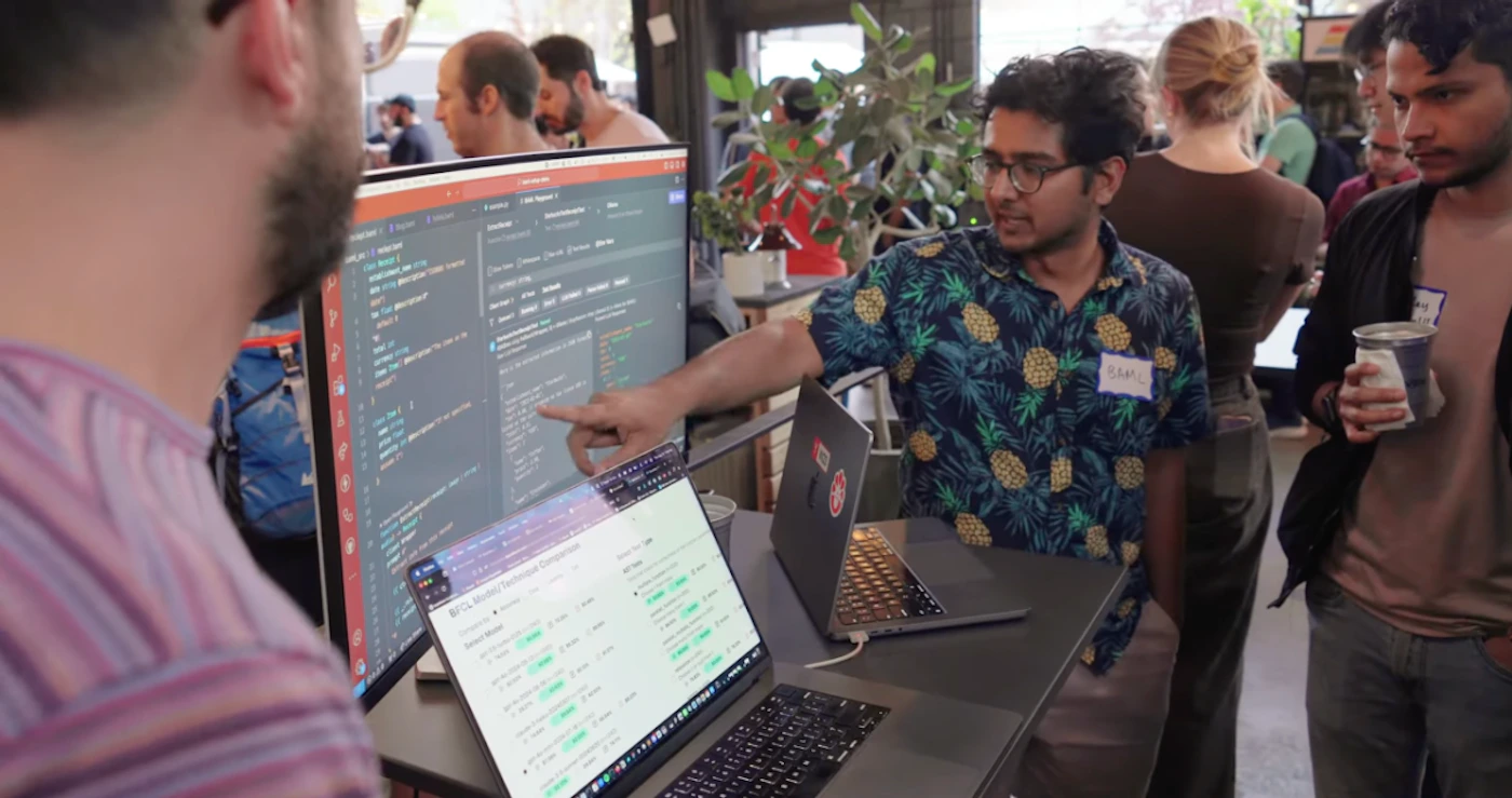 (Photo) A person points at a computer screen displaying code while others look on in a casual setting. people | indoor | computer screens, code, casual gathering | candid Note: The image is a real-life captured moment of people interacting with technology, making it a photograph. It's not staged or posed in a formal way.