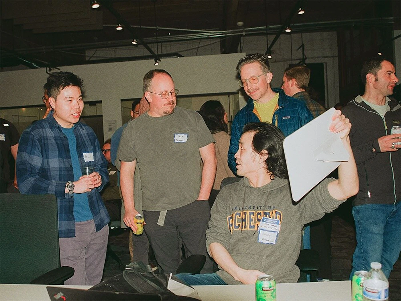 (Photo) This photo shows a group of people gathered indoors, likely at an event or meeting. Several individuals are engaged in conversation. Text: UNIVERSITY OF [unreadable] [unreadable] people | indoor | multiple people, name tags | candid | Colors: #778899, #000080, #000000 Note: The image captures a real-life scene with people, suggesting it's a photograph. There are no obvious signs of illustration, logo, banner or handwriting.